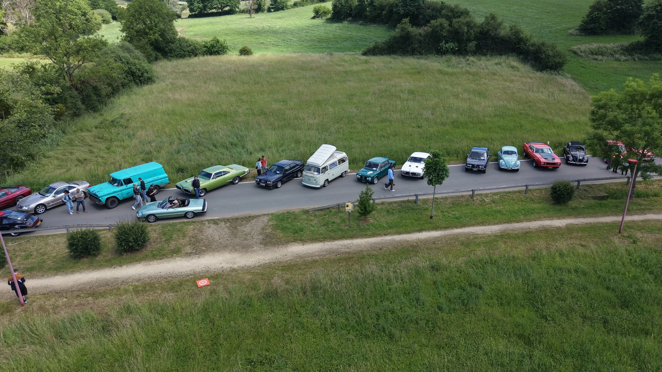 Aerial view of a row of classic and vintage cars parked along a rural road with people walking and taking photos, surrounded by green grass and trees.