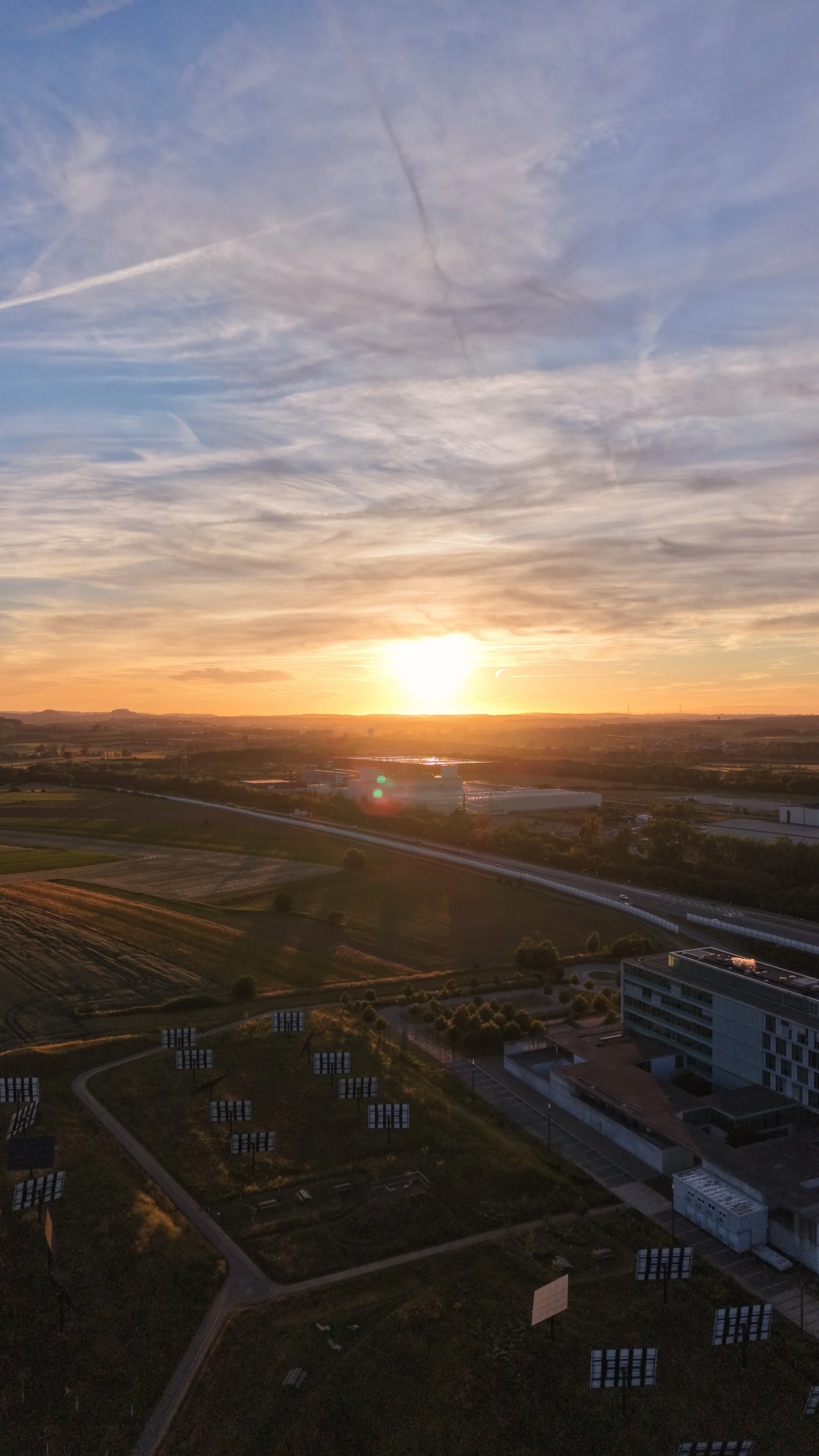 Sunset over a rural landscape with fields, roads, and some buildings, under a sky with scattered clouds and contrails.