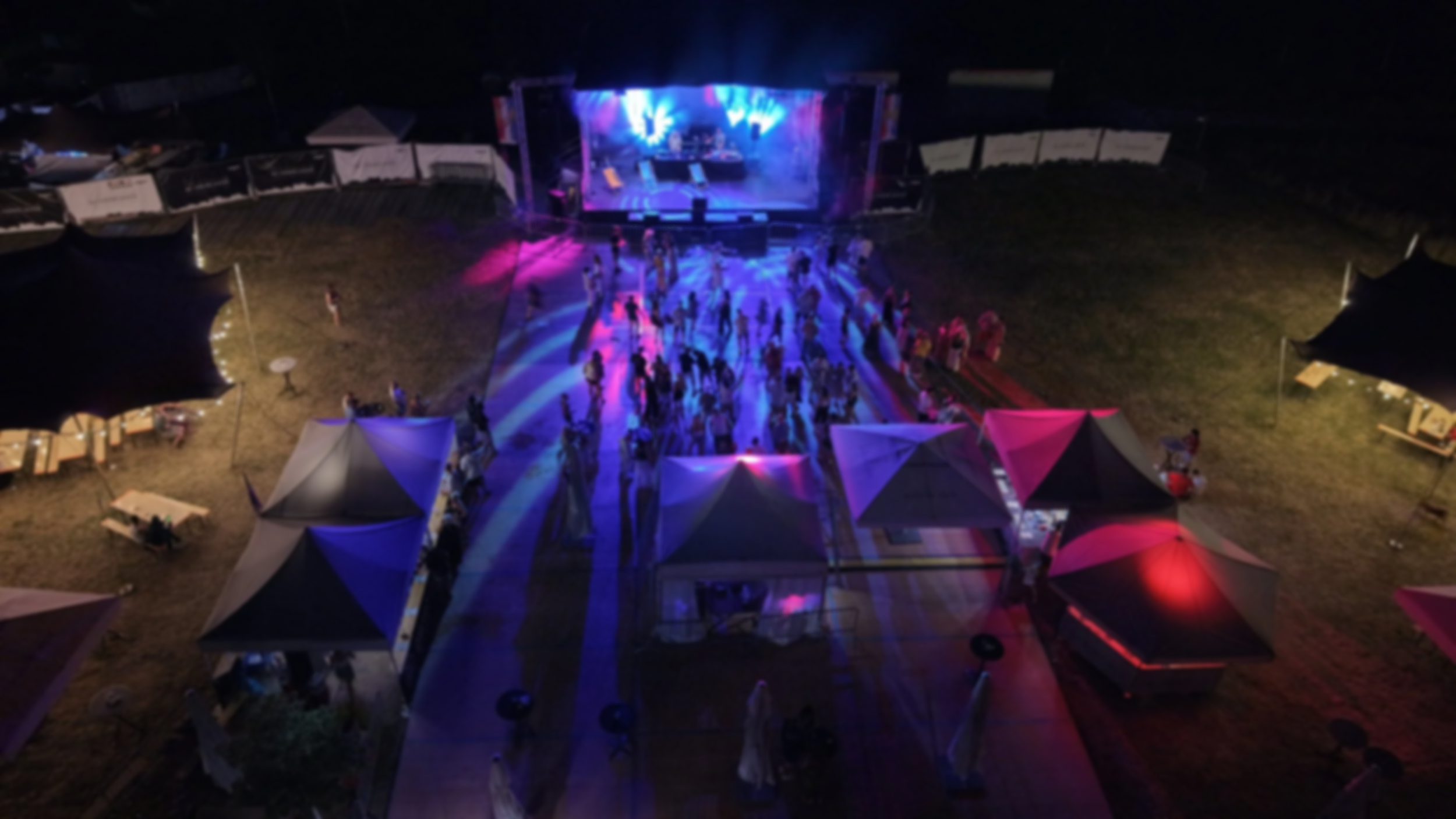 An outdoor night scene showing a stage with colorful lights, surrounded by tents, and a dance floor with people dancing.