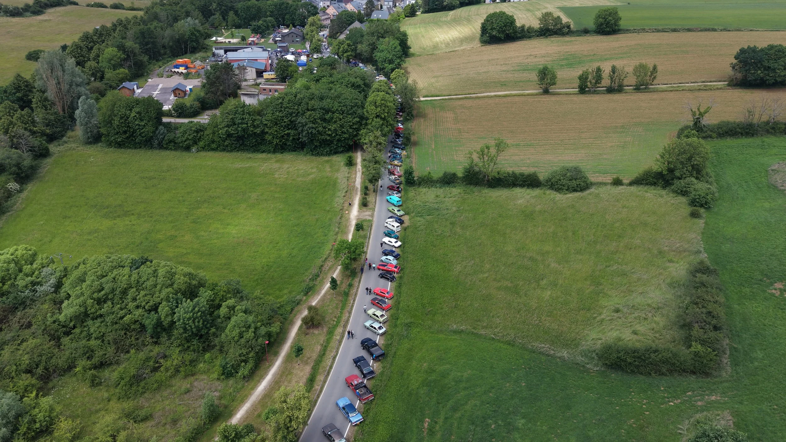 An aerial view of a rural landscape with fields, trees, and a narrow road running through the scene. Along the road, there are numerous parked cars and a small group of people walking.