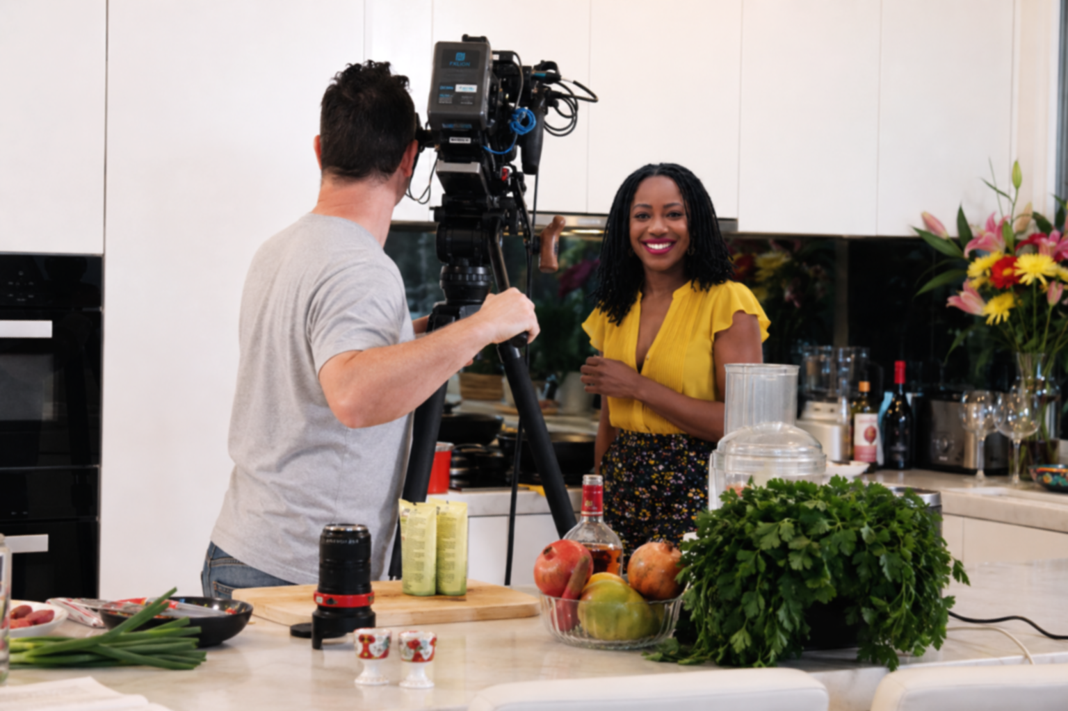 Chef May on a kitchen set, being filmed by a man with a camera.