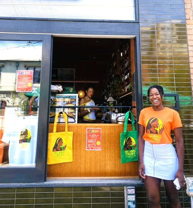 Chef May smiling and standing outside a restaurant window, wearing an Island crust orange t-shirt