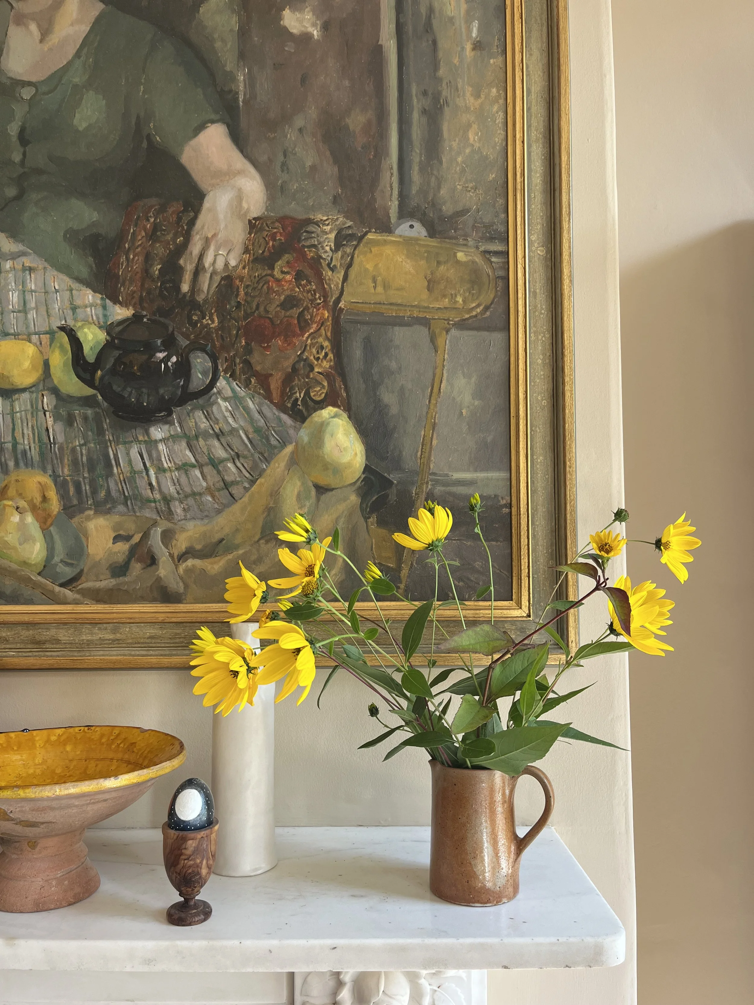 A ceramic vase with yellow flowers and green leaves on a white table, colorful still life painting in the background.