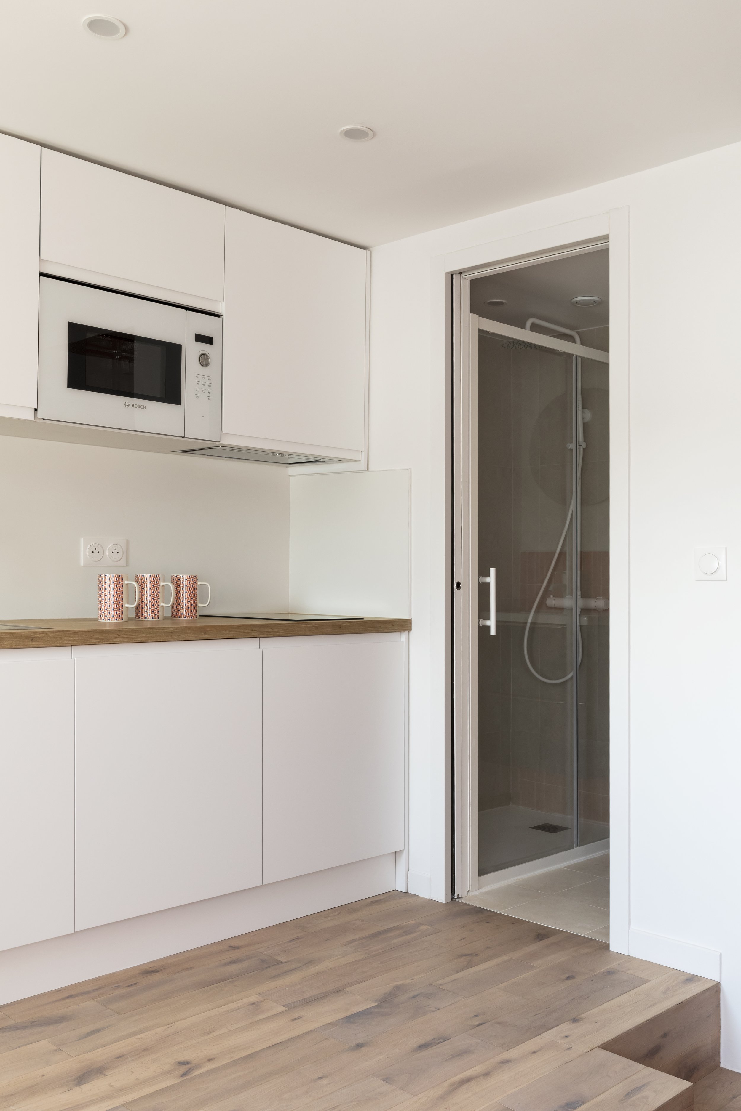 Une cuisine moderne avec des armoires blanches, un micro-ondes intégré, un plan de travail en bois, et une douche à l'italienne avec porte en verre
