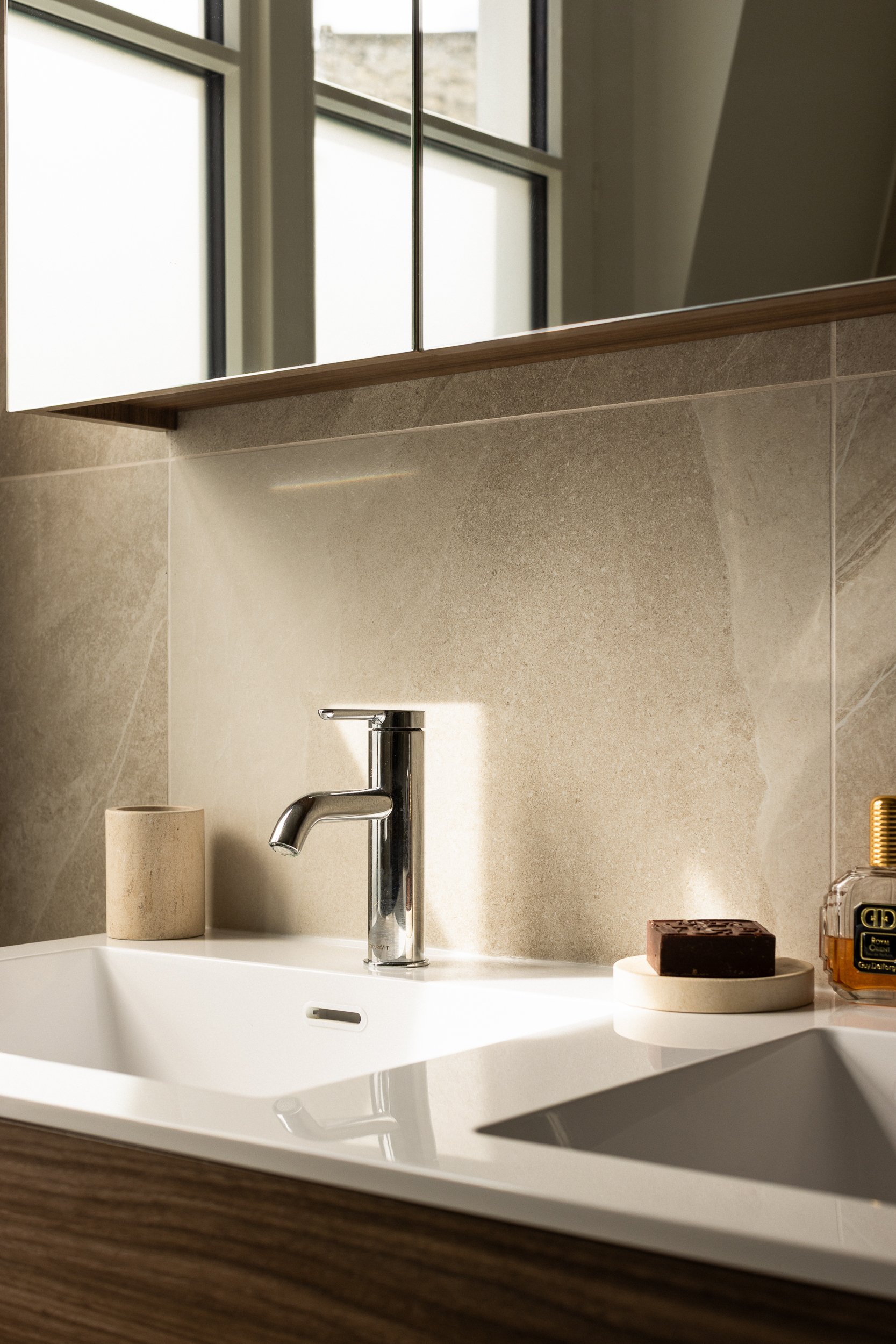 Vue partielle d'un lavabo avec un robinet en chrome, un porte-brosse à dents en bois, un savon en bloc sur un support rond et un flacon de savon liquide sur une surface en bois, dans une salle de bain avec un miroir.