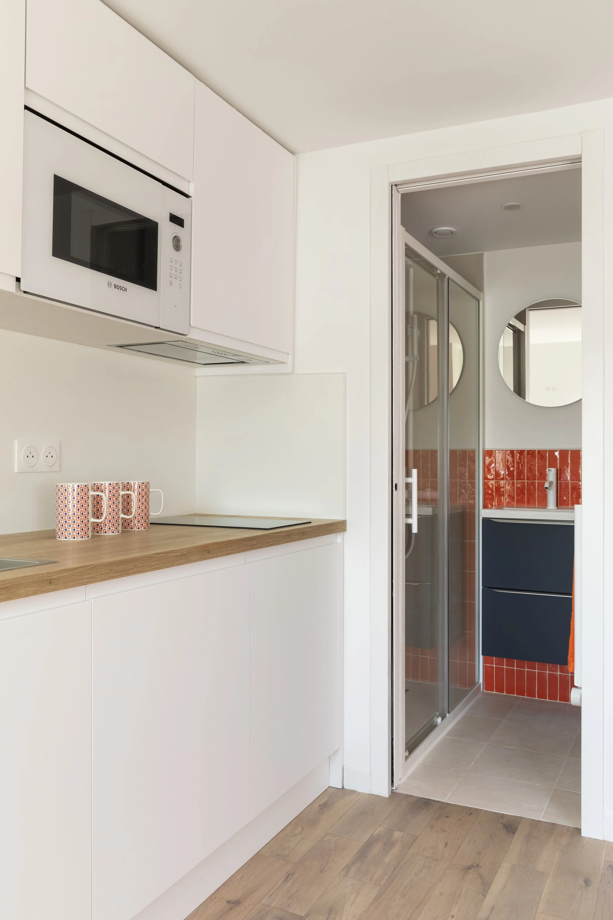 Une cuisine moderne avec des armoires blanches, une micro-ondes Bosch intégrée, un plan de travail en bois clair, et un espace salle de bain avec douche en verre et miroir rond, carrelage orange derrière le lavabo.