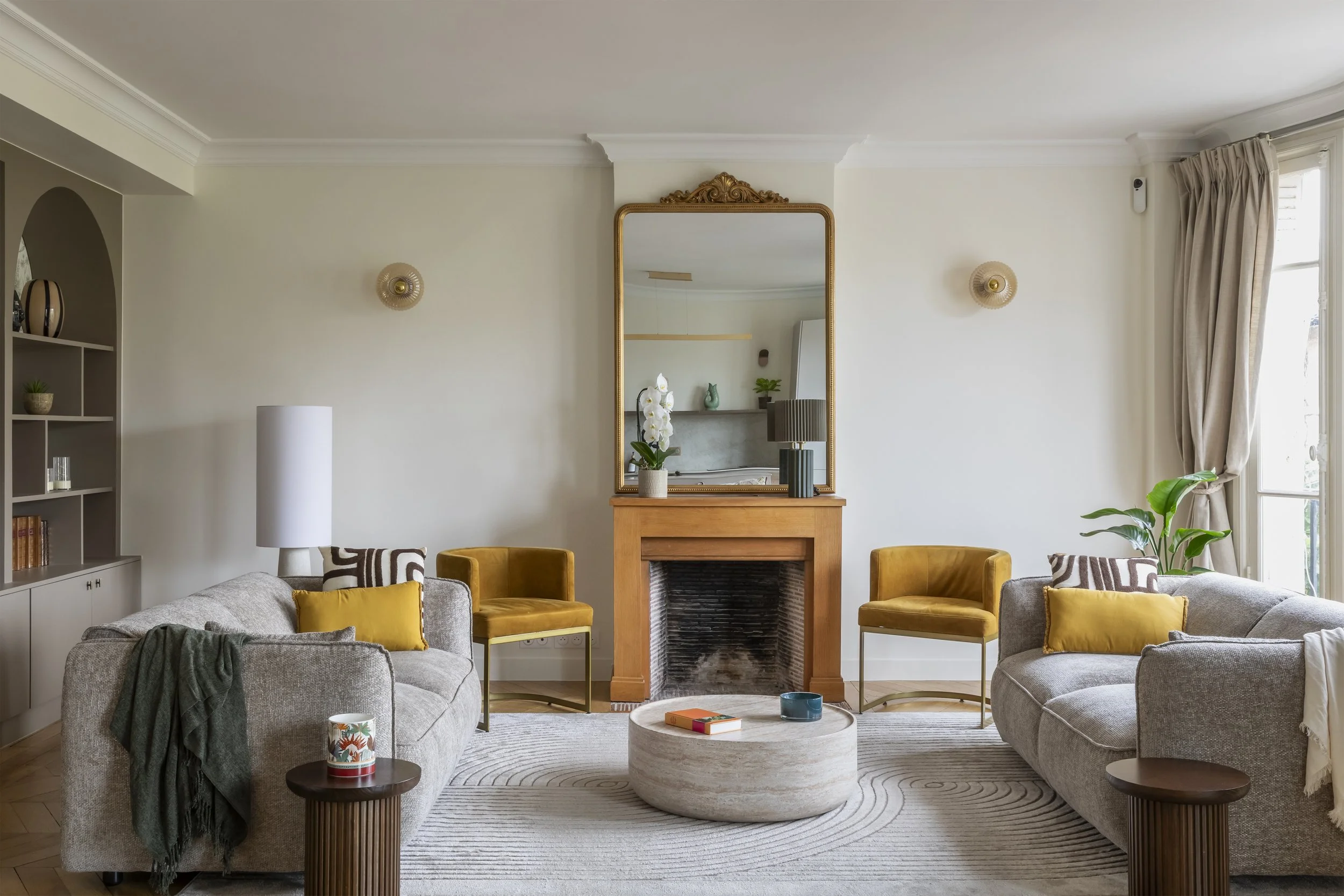 Salon avec cheminée, miroir doré, canapés gris, fauteuils jaunes, table basse en pierre, étagère avec décorations, lampes, et plantes, grande fenêtre avec rideaux