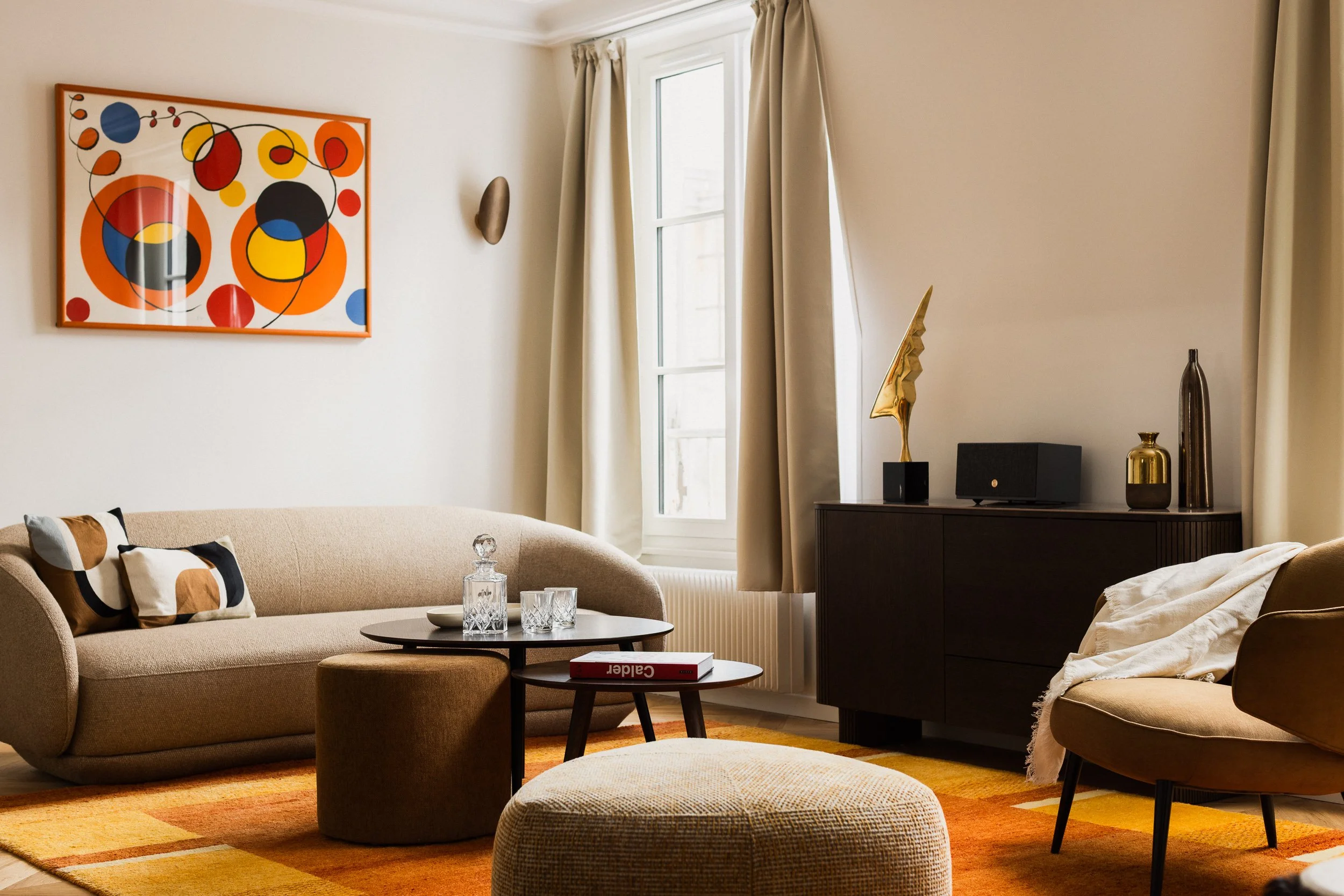 Salon contemporain dans appartement haussmannien avec canapé beige arrondi de la marque Boconcept modèle Bolsano, pouf triangulaire aux angles arrondis de la marque Boconcept modèle Bermuda, tableau abstrait coloré sur le mur, tables basses rondes gi