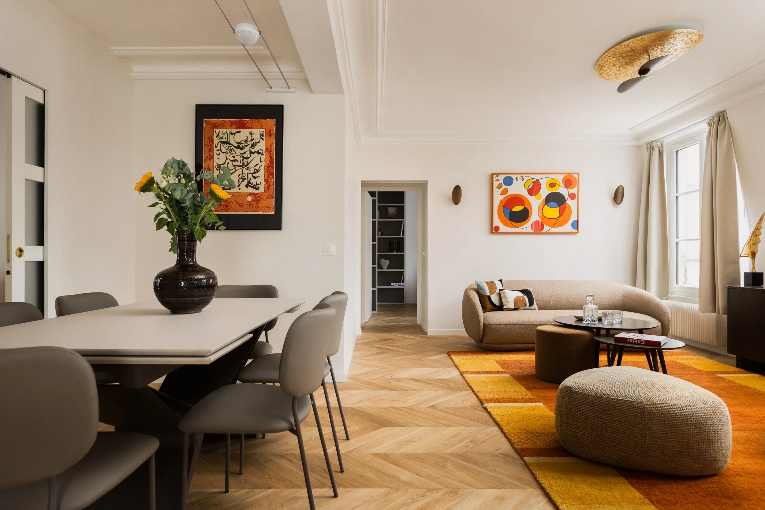 Pièce à vivre contemporaine dans appartement haussmannien avec table à manger extensible de marque Italienne, canapé beige arrondi de la marque Boconcept modèle Bolsano, pouf triangulaire aux angles arrondis de la marque Boconcept modèle Bermuda, un 