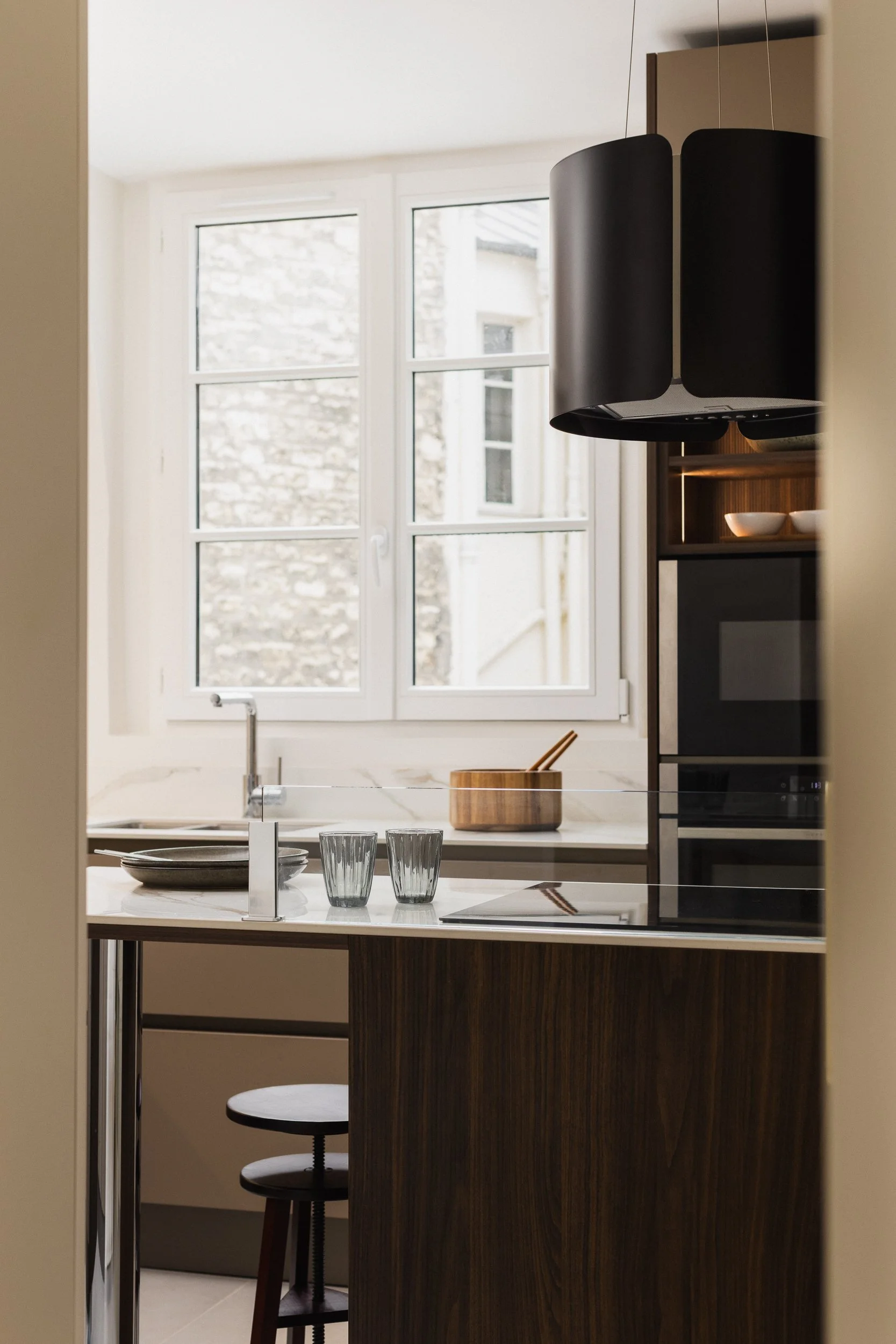 Vue d'une cuisine moderne contemporaine dans un intérieur Haussmannien à travers une ouverture, avec une fenêtre en arrière-plan, une table avec des assiettes, des verres et une boîte à couverts en bois, et des meubles en bois foncé et clair. Une hot
