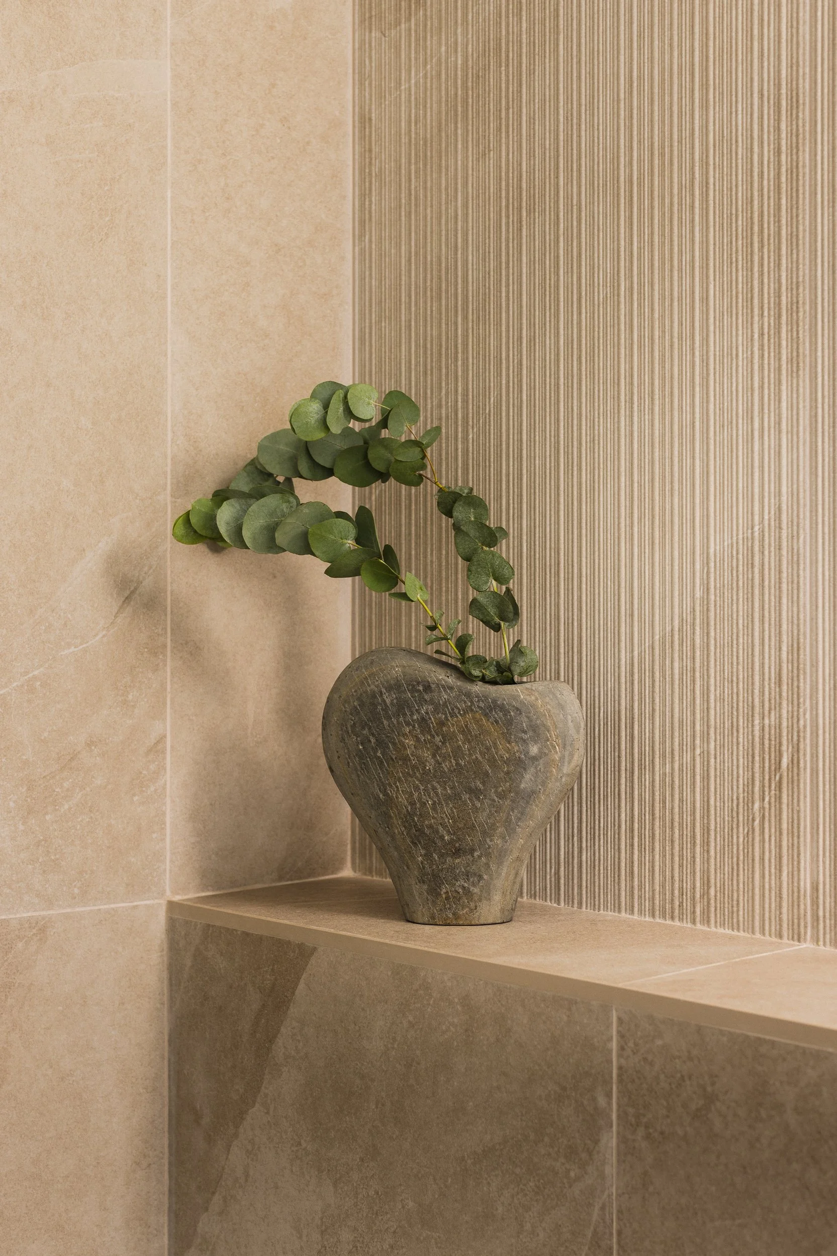 Vase en pierre de la marque BoConcept modèle Burly contenant un feuillage branches eucalyptus vert, posé sur une niche dans un espace intérieur de salle de bain, avec murs en carrelage carreaux de céramique beige de la marque Italgraniti collection S