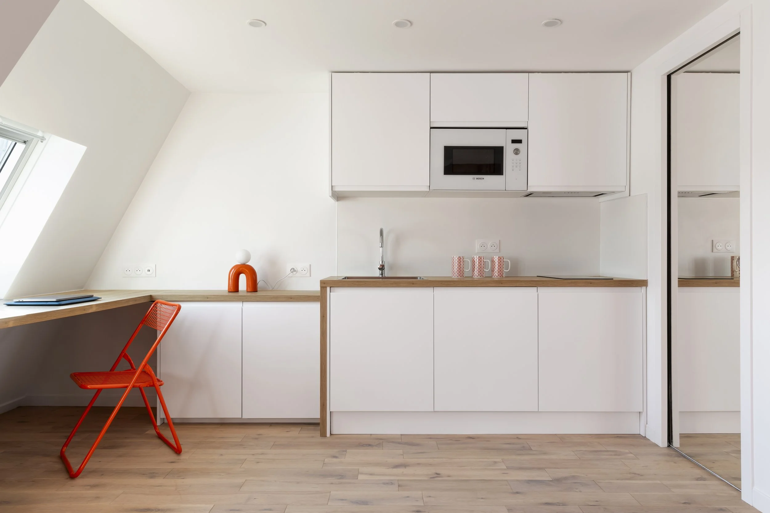 Cuisine moderne avec comptoir en bois, armoires blanches, micro-ondes Bosch intégré, fenêtres inclinées, sol en bois clair, une chaise rouge en métal et des tasses sur le comptoir.