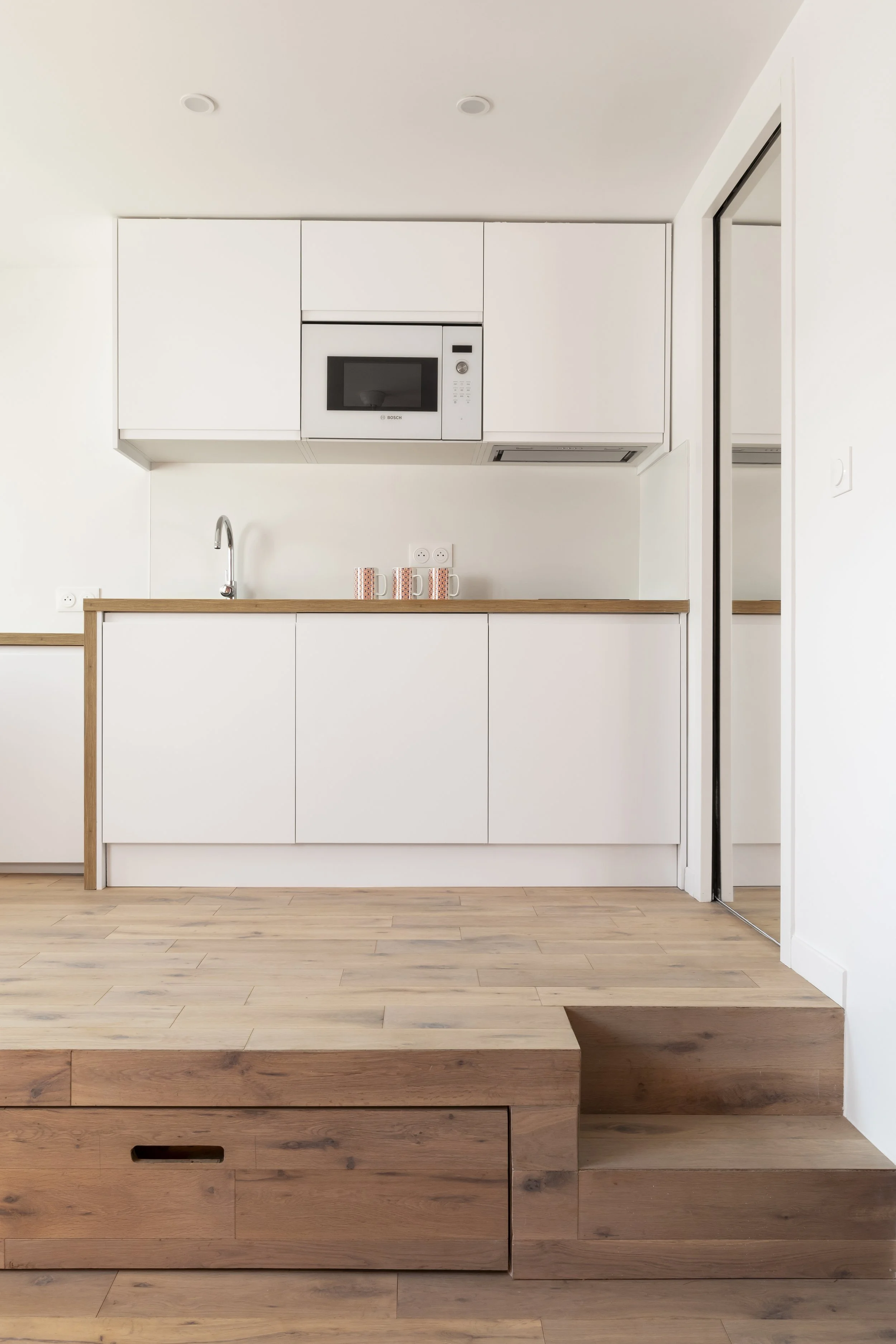 Cuisine moderne avec comptoir en bois clair, armoires blanches, micro-ondes intégré, évier en acier inoxydable, sol en bois clair, pas de décoration visible.
