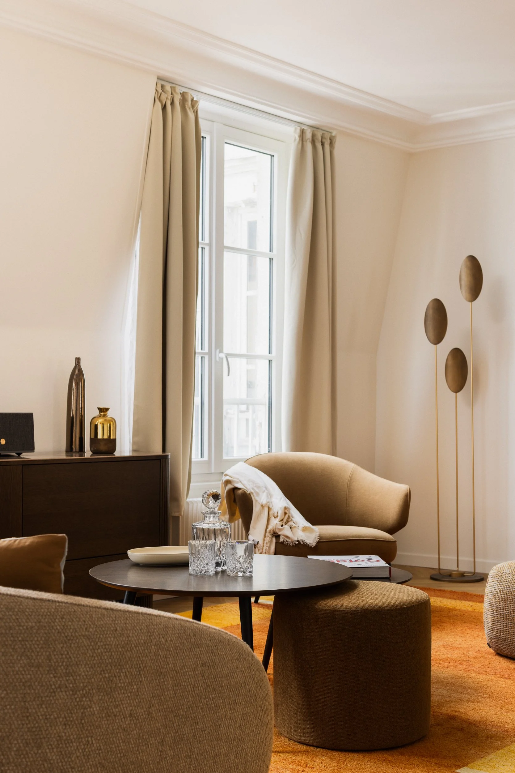 Salon contemporain dans appartement haussmannien avec table à manger extensible, lampadaire d'inspiration Art Déco, tables basses rondes gigognes en bois, pouf d'appoint, objets décoratifs en or et brun, grande fenêtre laissant entrer la lumière natu