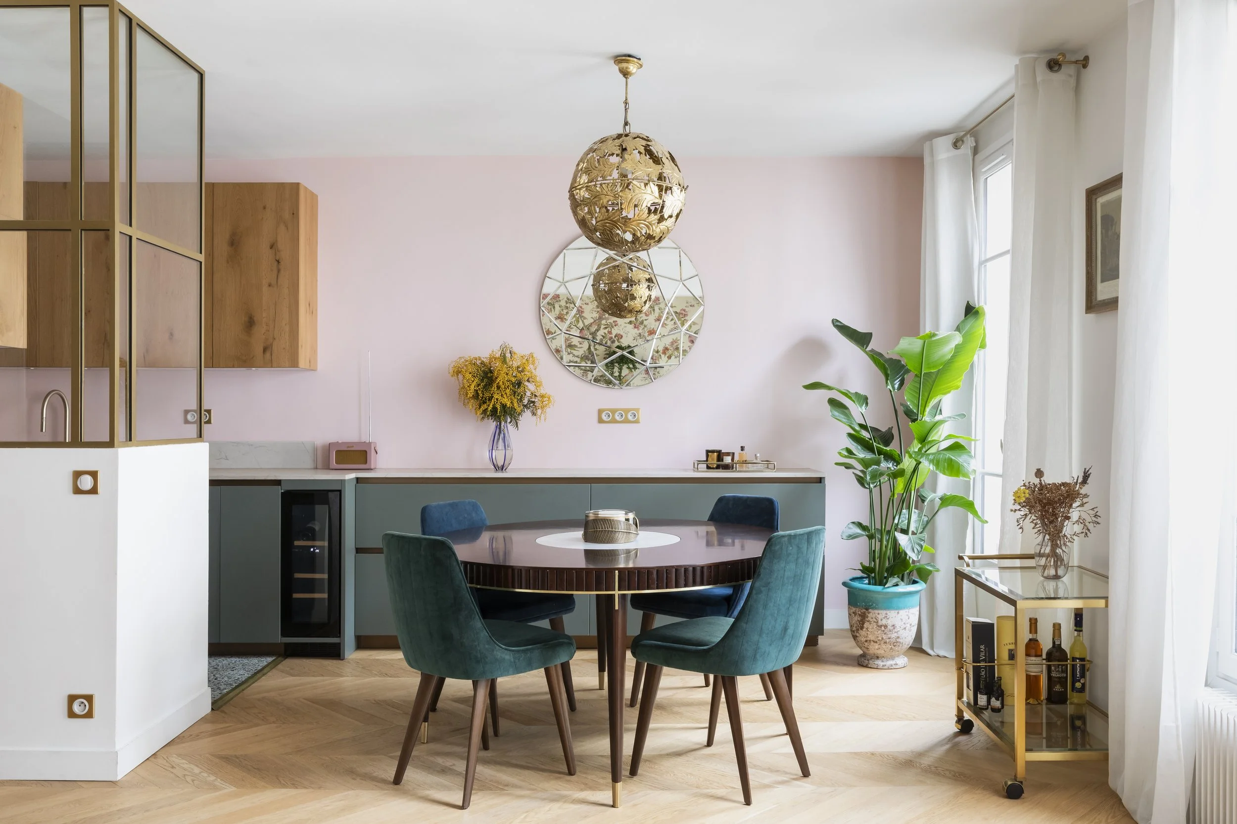 Intérieur d'un salon avec un meuble de cuisine en fond, un miroir rond, un lustre en forme de sphere en or, une grande plante verte, une table ronde avec des chaises en velours vert, et un meuble en métal avec des bouteilles et des livres.