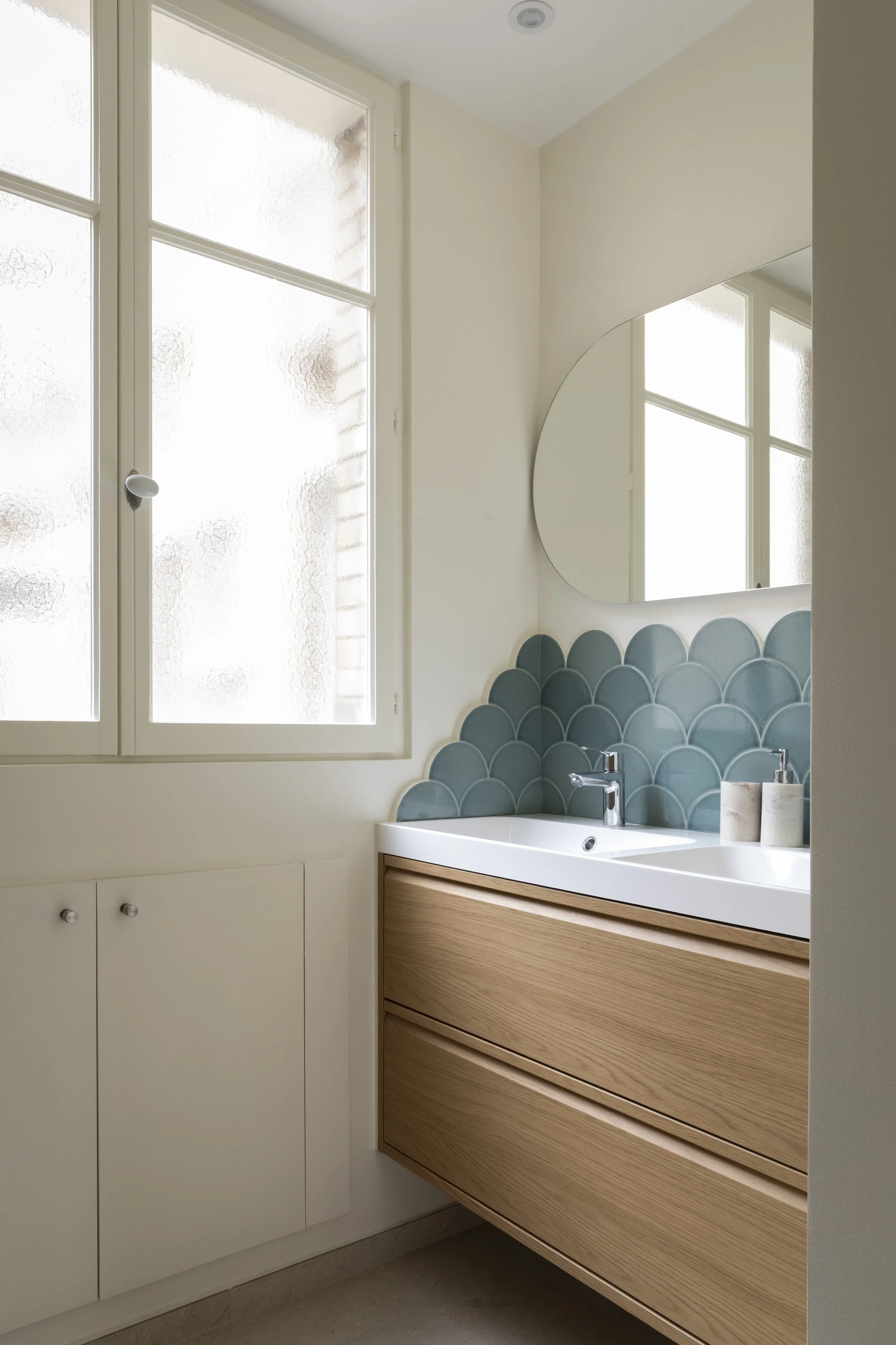 Vue partielle d'une salle de bain avec une fenêtre, un miroir rond, un lavabo blanc, un meuble en bois clair avec tiroirs, et un mur avec des carreaux en forme d'écailles bleues.