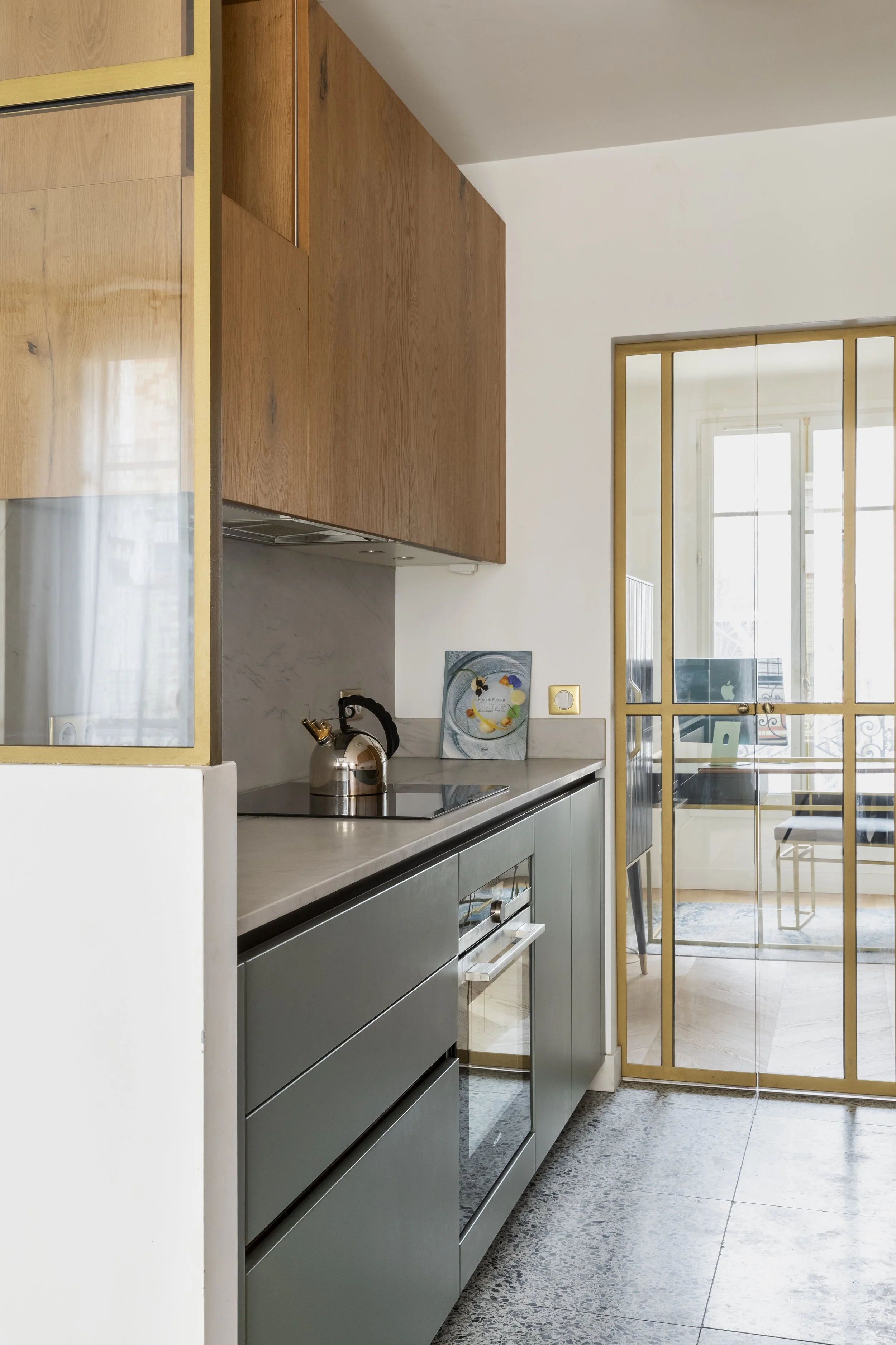 Cuisine moderne avec meubles gris, étagère en bois, porte vitrées avec cadre en bois, et éléments décoratifs tels qu'une peinture, une théière, et un ordinateur visible à l'arrière.