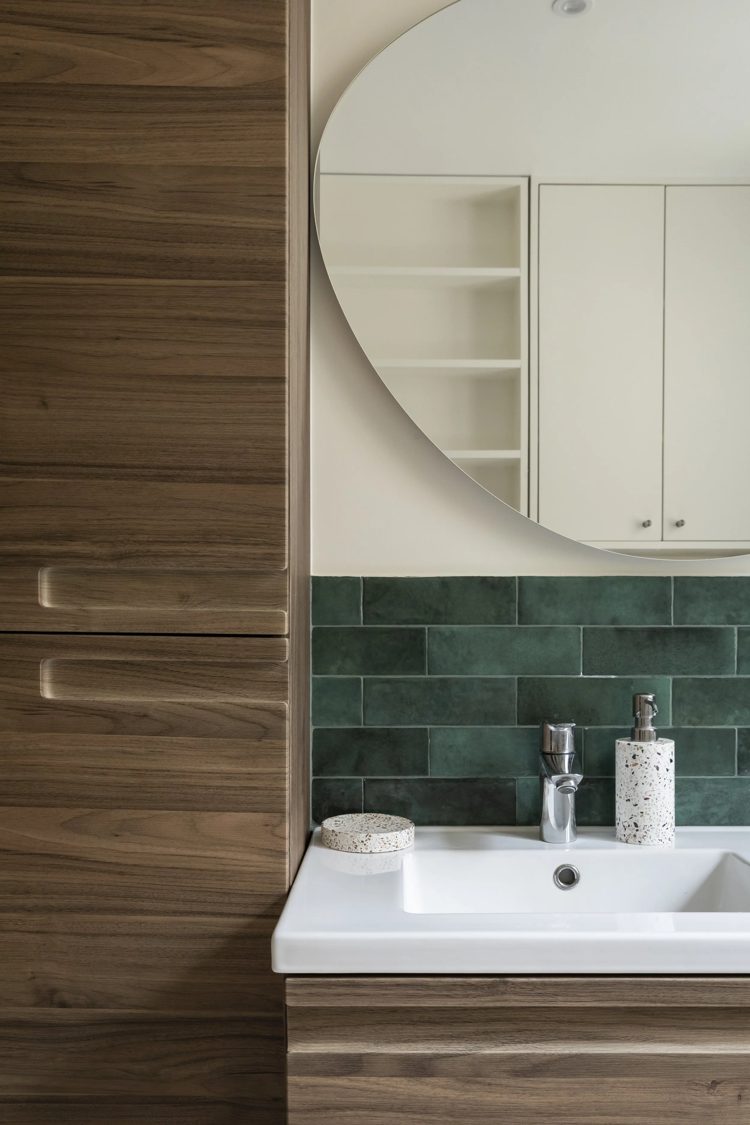 Un lavabo blanc avec un robinet en métal, un porte-savon à broderie, et un distributeur de savon dans une salle de bain moderne avec un mur en carreaux verts et un meuble en bois.