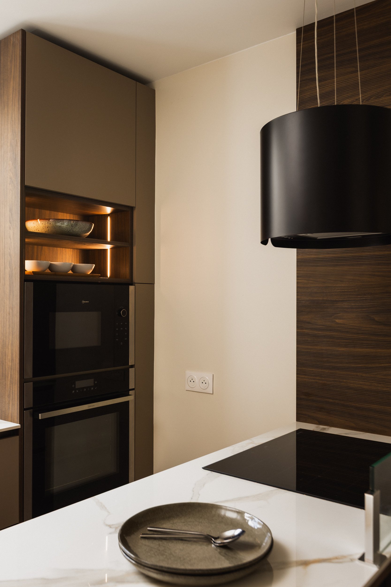Cuisine moderne contemporaine avec four intégré, plaque de cuisson noire et hotte noire suspendue, plan de travail en céramique marbre blanc veiné, et une assiette avec des couverts en gros plan.