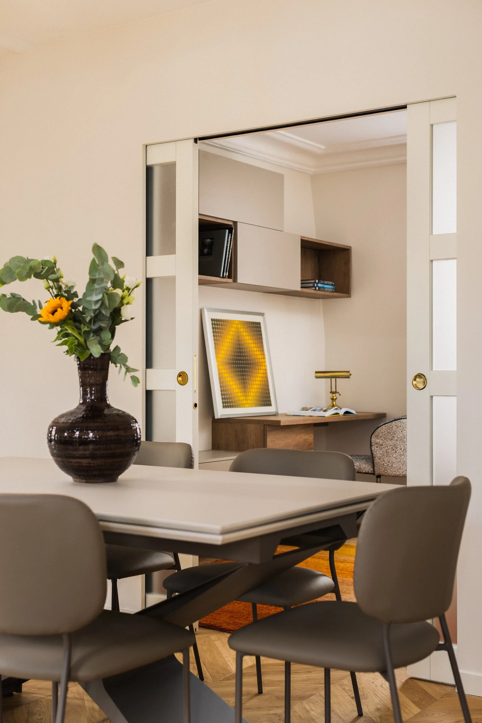 Une salle à manger contemporaine dans un intérieur Haussmannien avec une table en métal extensible entourée de 6 chaises en métal et cuir. Au centre de la table, un vase noir avec des fleurs vertes et une fleur jaune. À l'arrière d'une porte à galand