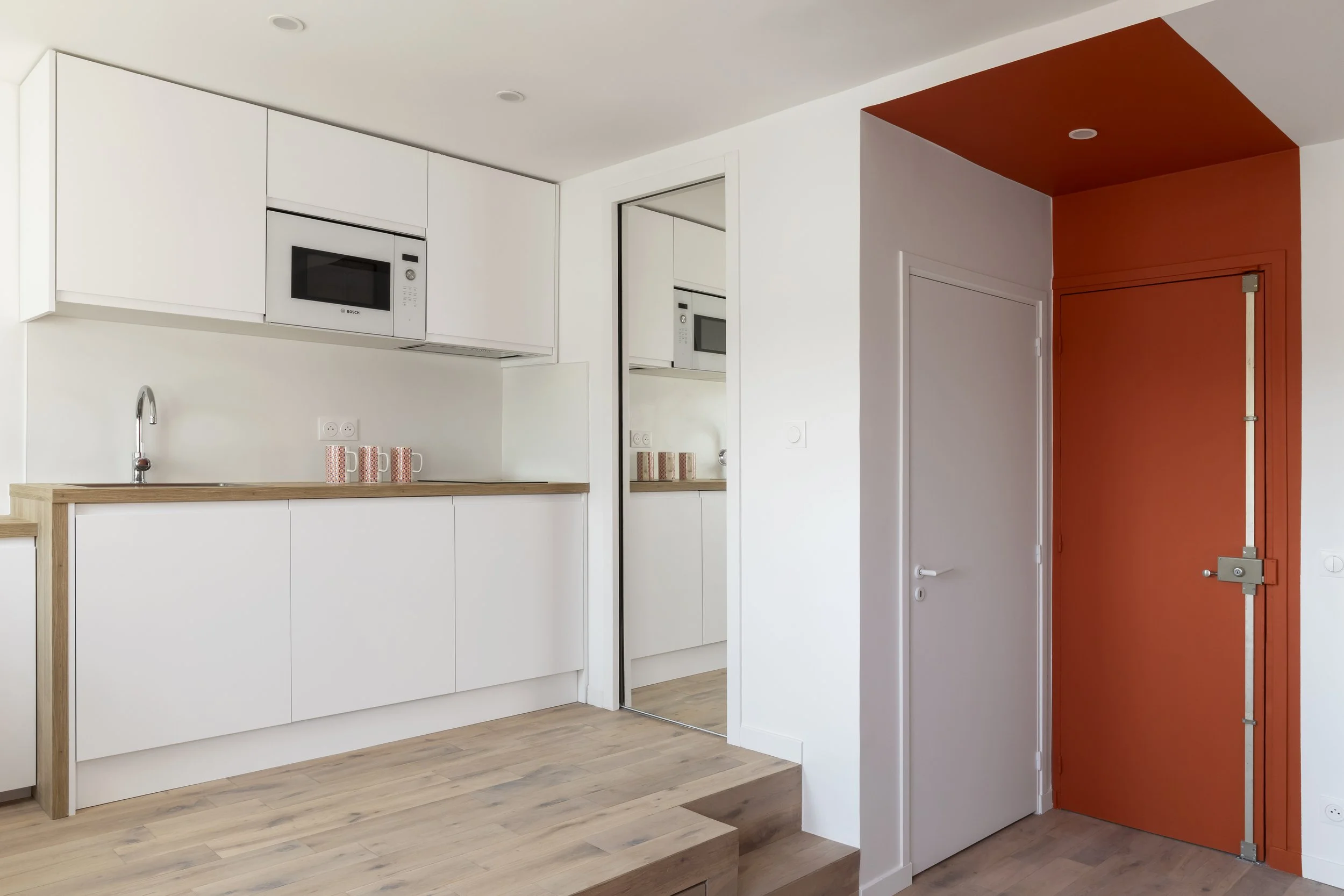 Cuisine moderne avec armoires blanches, micro-ondes intégré, comptoir en bois, mur blanc, porte rouge avec système de fermeture par rouleau, sol en bois clair.