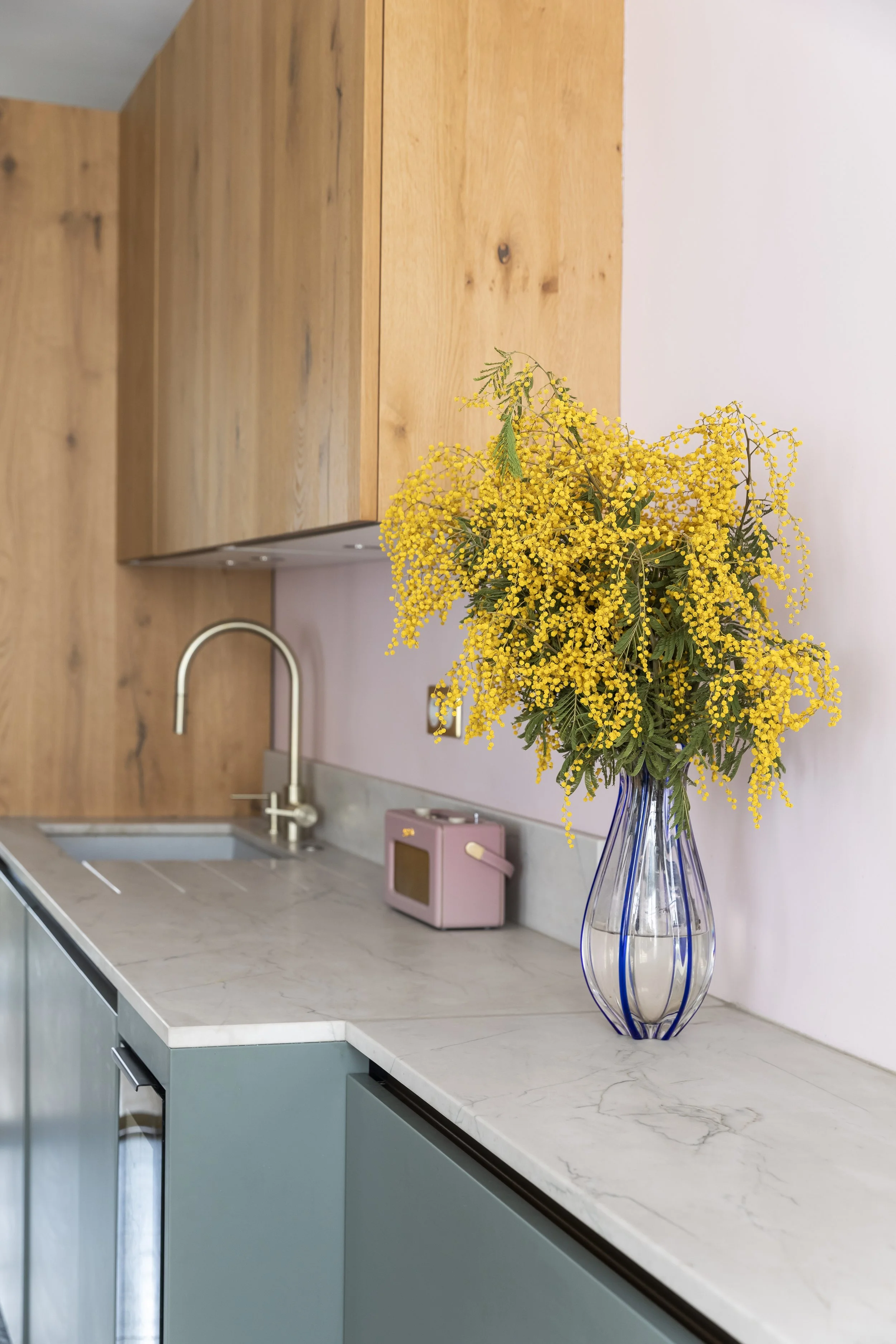 Vase en verre transparent avec des fleurs jaunes dans une cuisine moderne avec plan de travail en marbre blanc, armoires en bois et un robinet en métal.