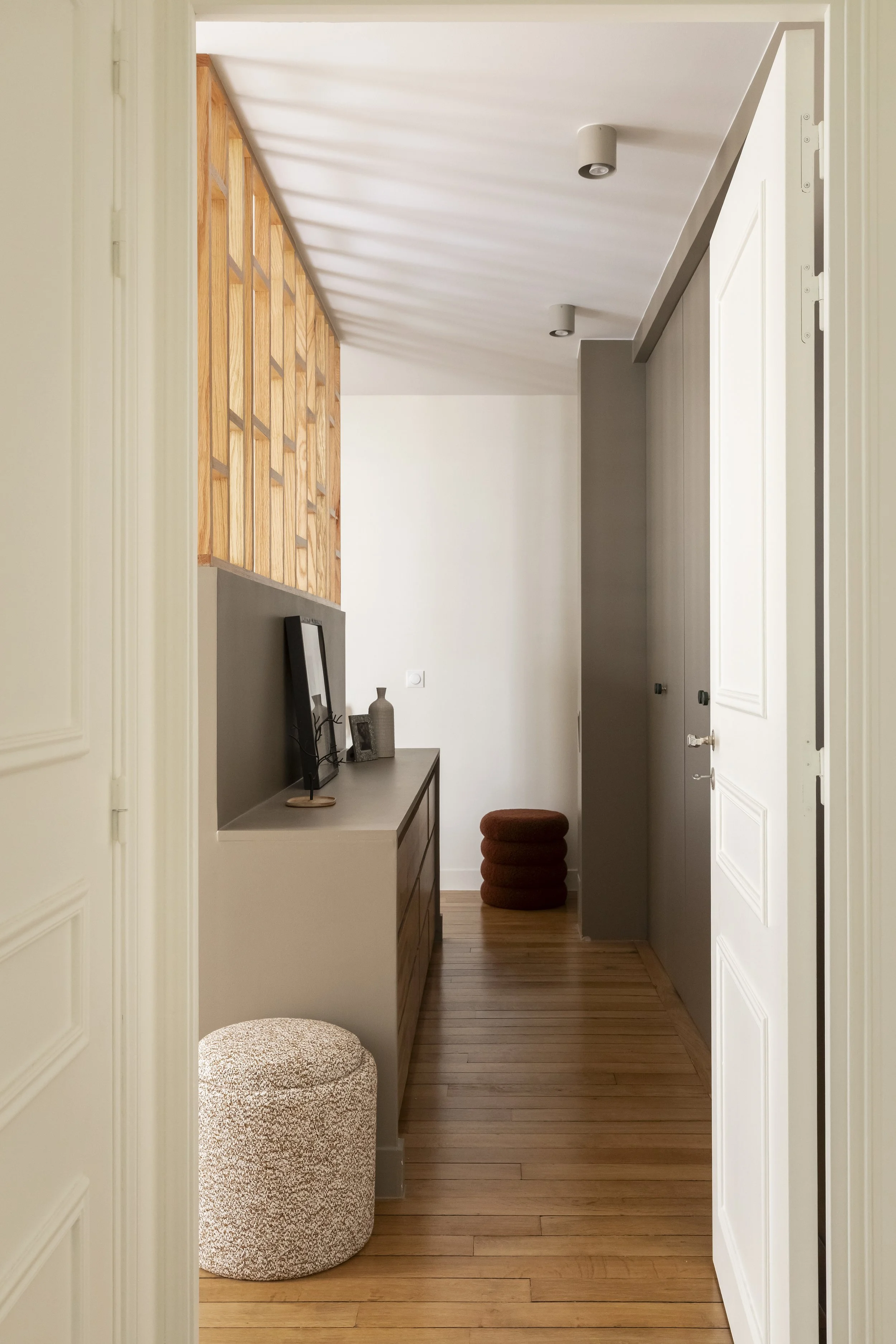 Un couloir intérieur avec un sol en bois clair, une console avec des objets décoratifs, et un mur avec une cloison en bois ajourée. Il y a aussi un pouf rond en tissu veiné marron et un rideau blanc.