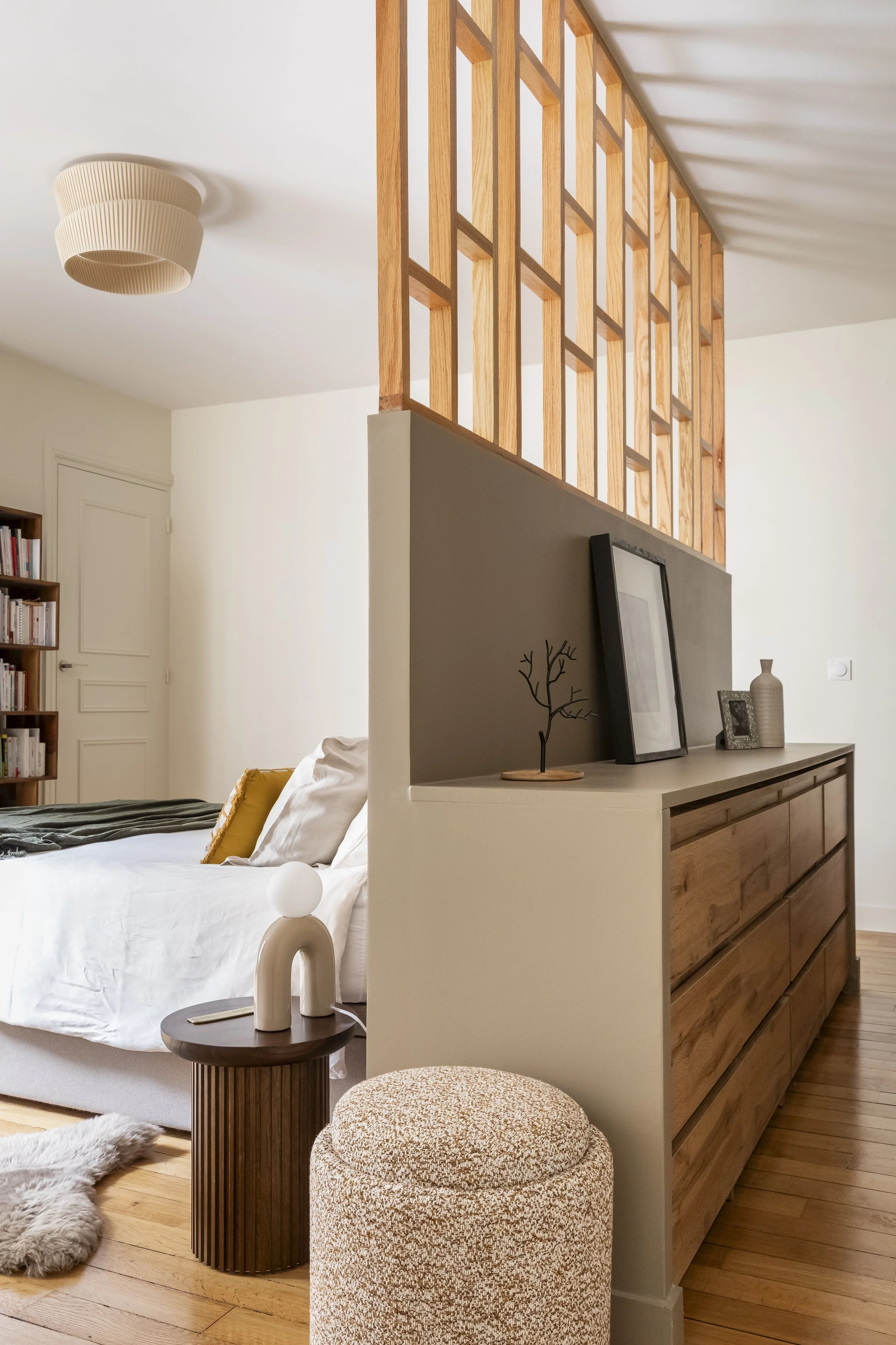 Une chambre à coucher moderne avec un lit blanc, des oreillers et une couverture sombre. À côté du lit, une petite table en bois avec une lampe décorative en forme de poignée de porte blanche. Dans l'espace séparé par une barrière en bois avec une gr