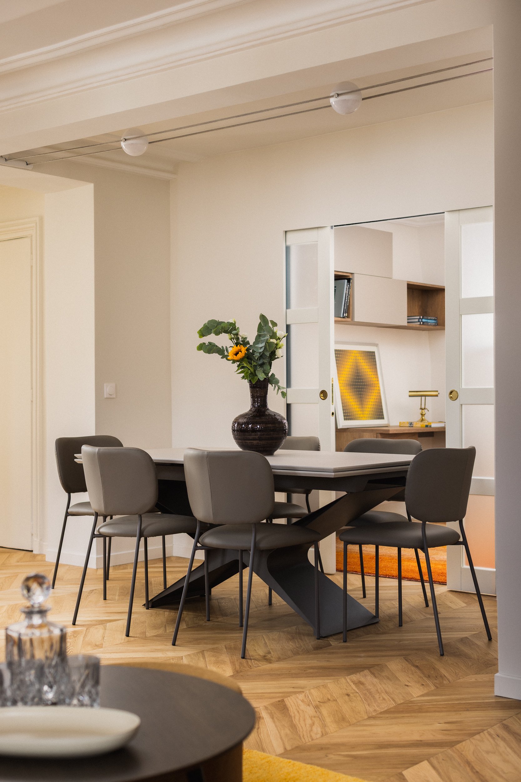 Une salle à manger contemporaine dans un intérieur Haussmannien avec une table en métal extensible entourée de 6 chaises en métal et cuir. Au centre de la table, un vase noir avec des fleurs vertes et une fleur jaune. À l'arrière d'une porte à galand