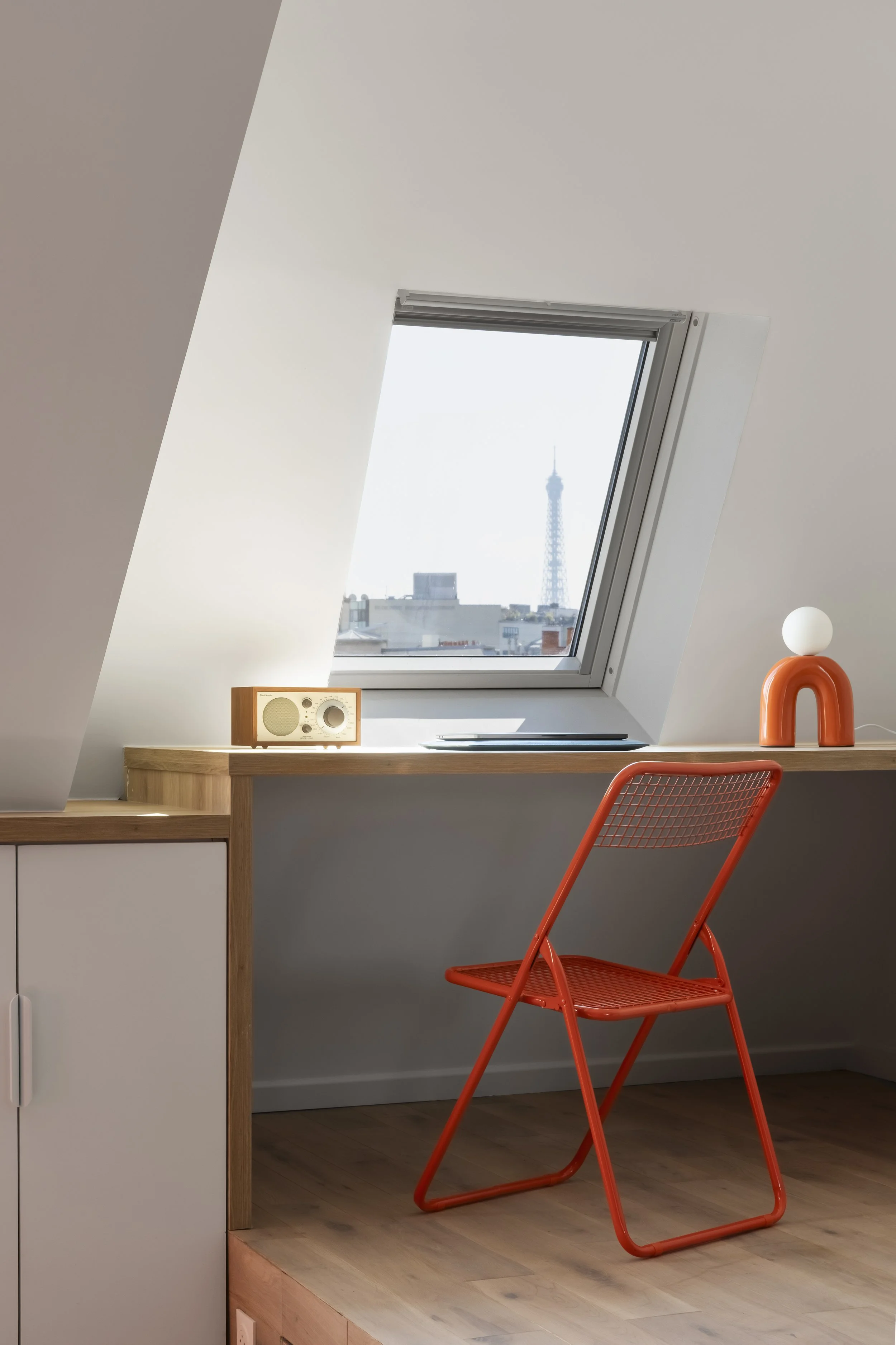 Une pièce avec un bureau en bois clair, une chaise orange en métal, une fenêtre de toit donnant sur Paris avec la Tour Eiffel en arrière-plan, un ventilateur en bois et un objet de décoration orange avec une boule blanche.