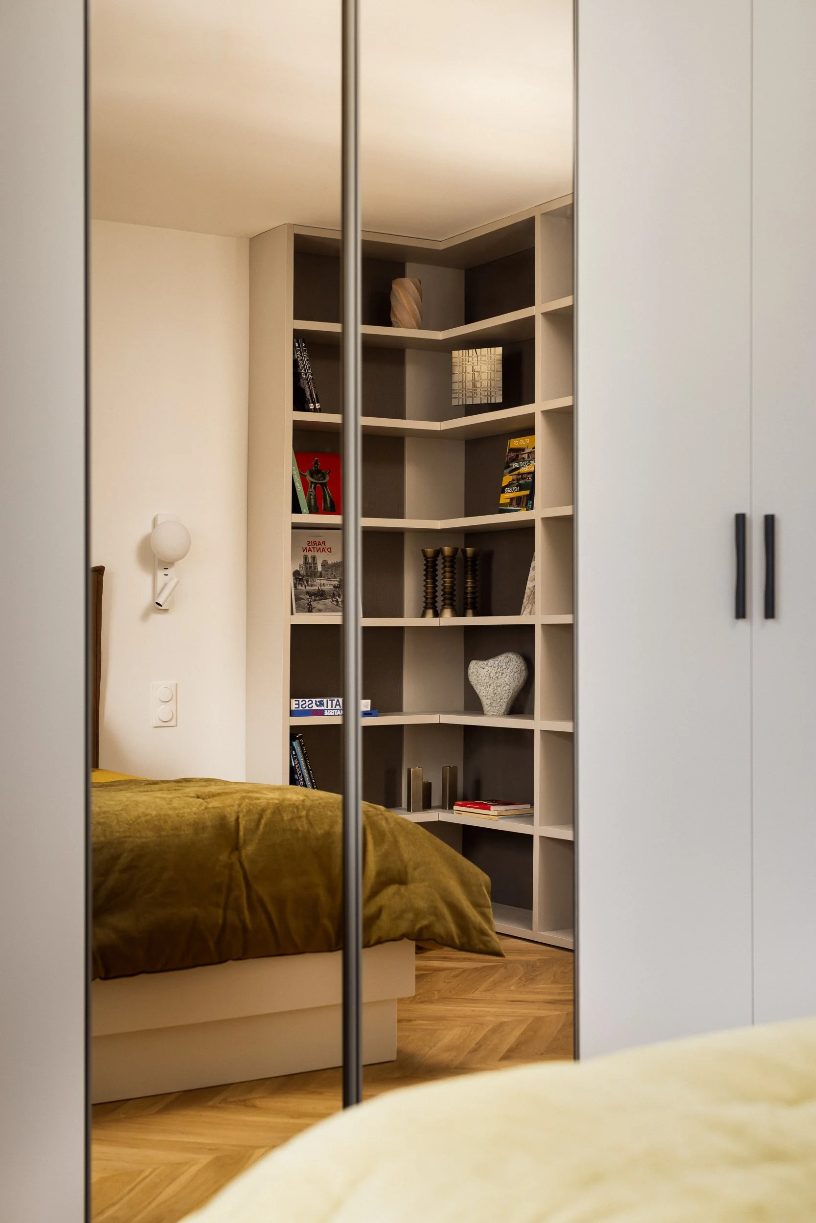 Une chambre parentale d'un intérieur Haussmanien vue à travers un miroir, montrant un lit avec boutis couvre lit de couleur bronze de la marque Bonsoirs, une bibliothèque toute hauteur sur mesure remplie de livres et de décorations, avec un sol en pa