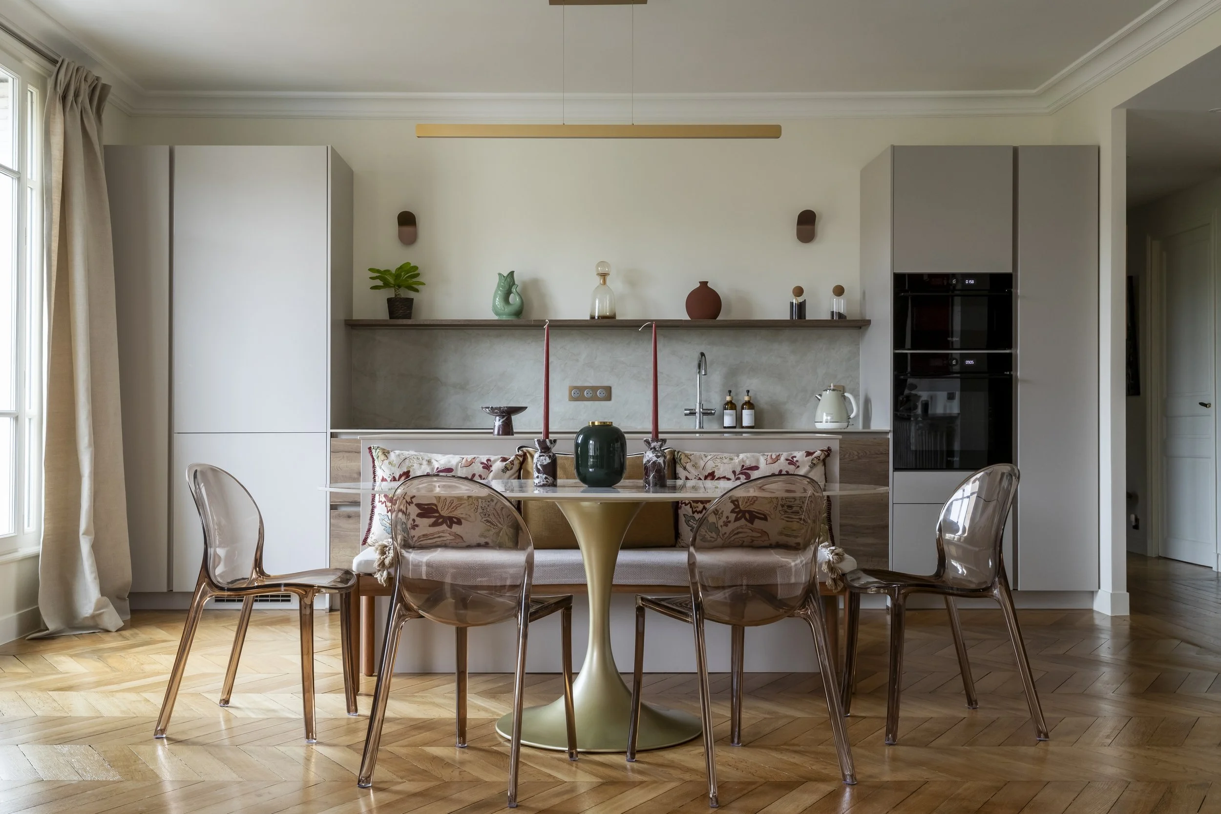 Une cuisine moderne contemporaine avec îlot central installée dans la pièce à vivre d'un appartement familial dans l'ancien, dans la avec une table à manger au pied circulaire doré et plateau ovale en céramique entourée de chaises en plastique polypr
