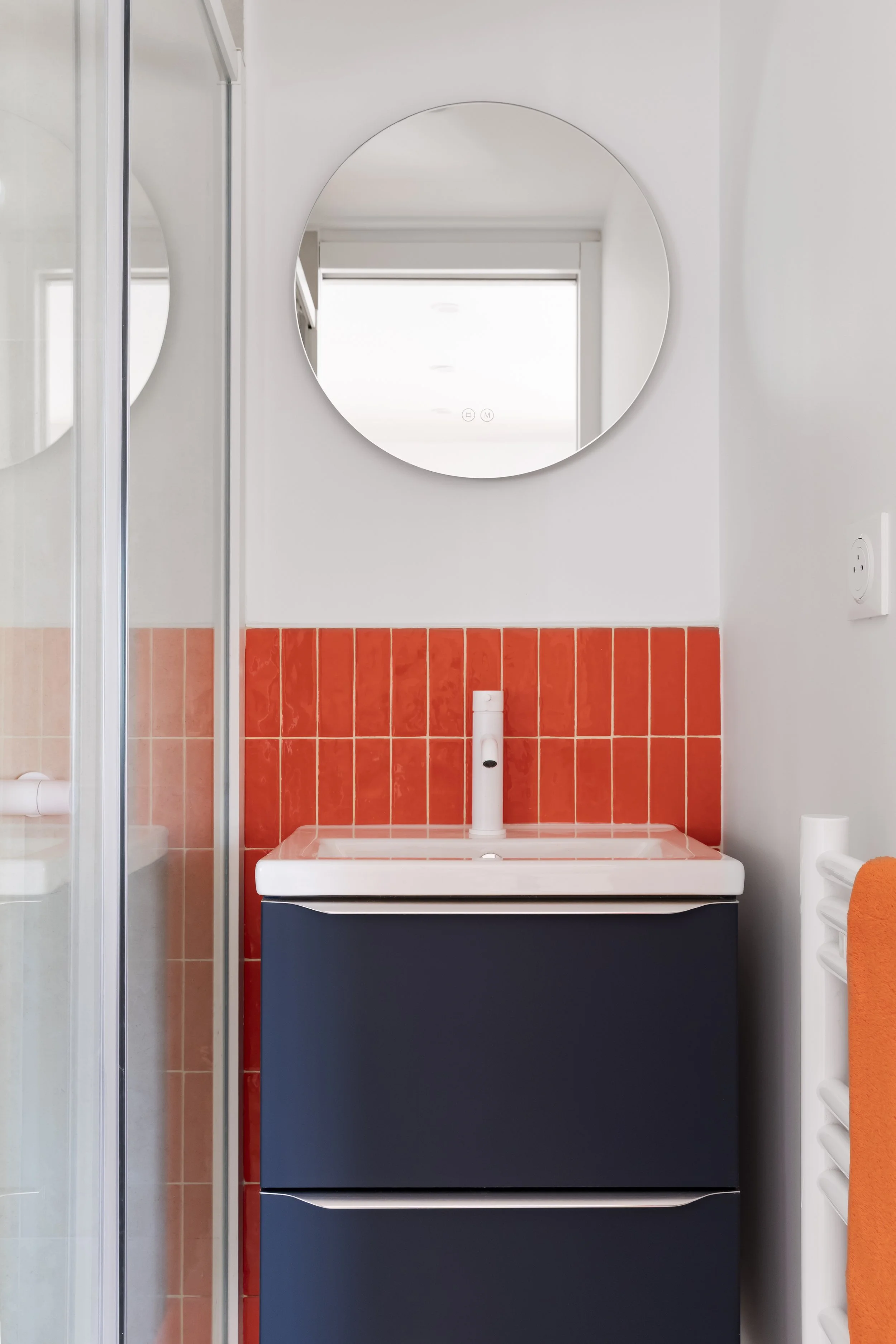 Un lavabo blanc avec un meuble bleu foncé en dessous, un miroir rond au-dessus, et un mur partiellement recouvert de carreaux orange, dans une salle de bain moderne.