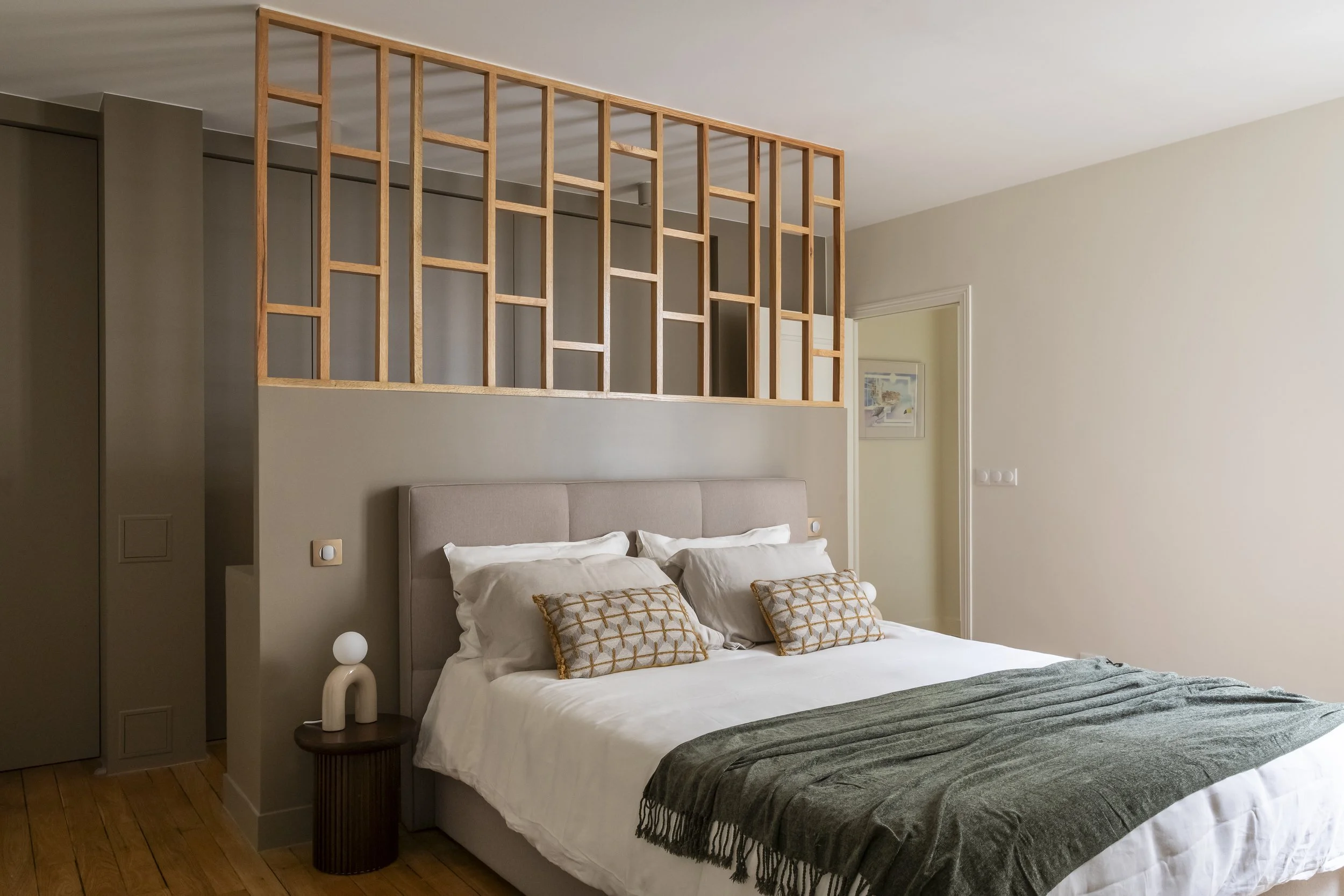 Chambre avec un lit, plusieurs oreillers avec des housses motifs, un tapis gris foncé, une tête de lit beige, et une structure en bois devant la tête de lit séparant la chambre d'une autre zone.