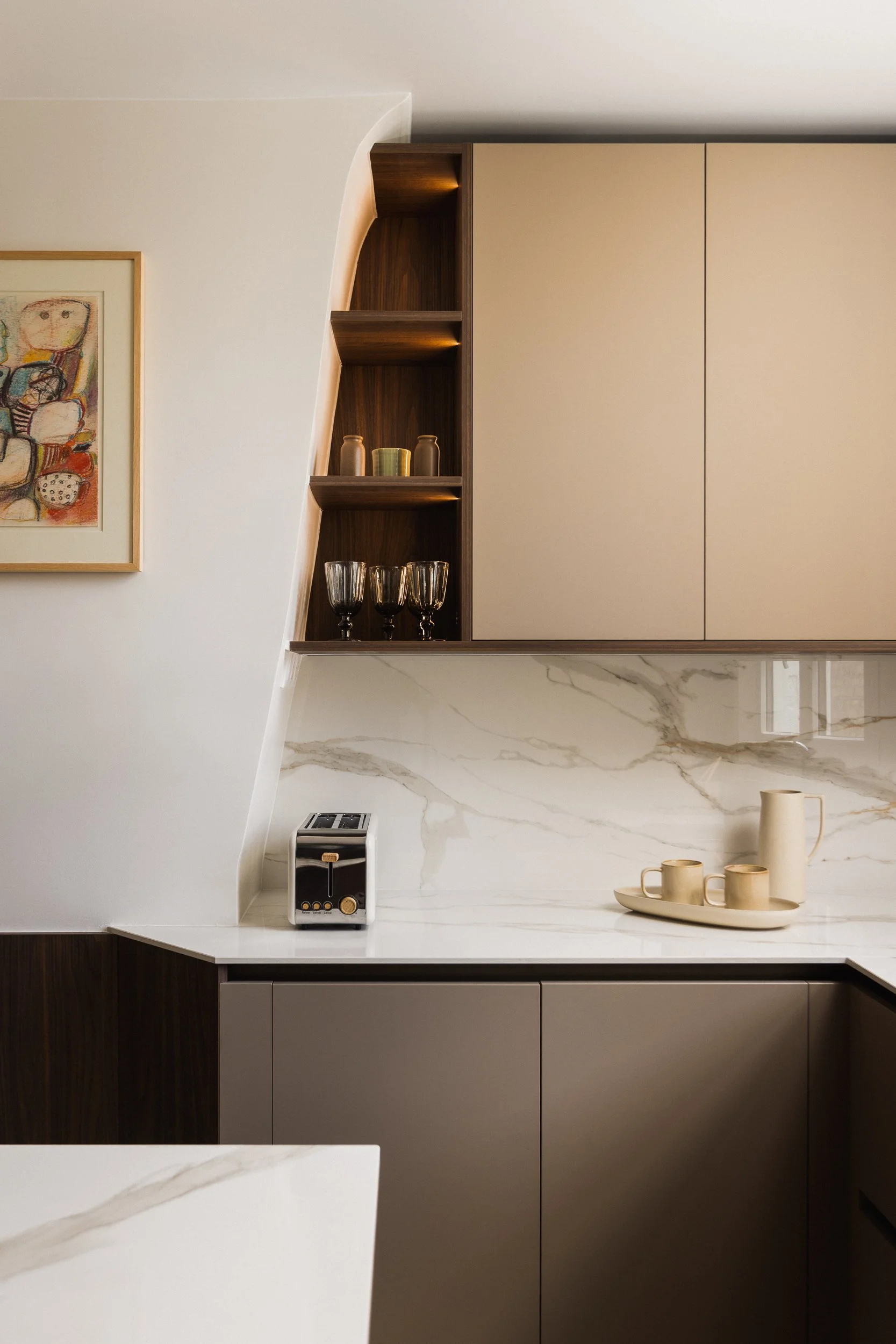 Une cuisine moderne contemporaines avec des placards beiges et taupe, des étagères niches en bois , un comptoir en céramique marbre blanc avec des veines grises, et des objets décoratifs comme des verres, des vases et des mugs dans un plateau beige.