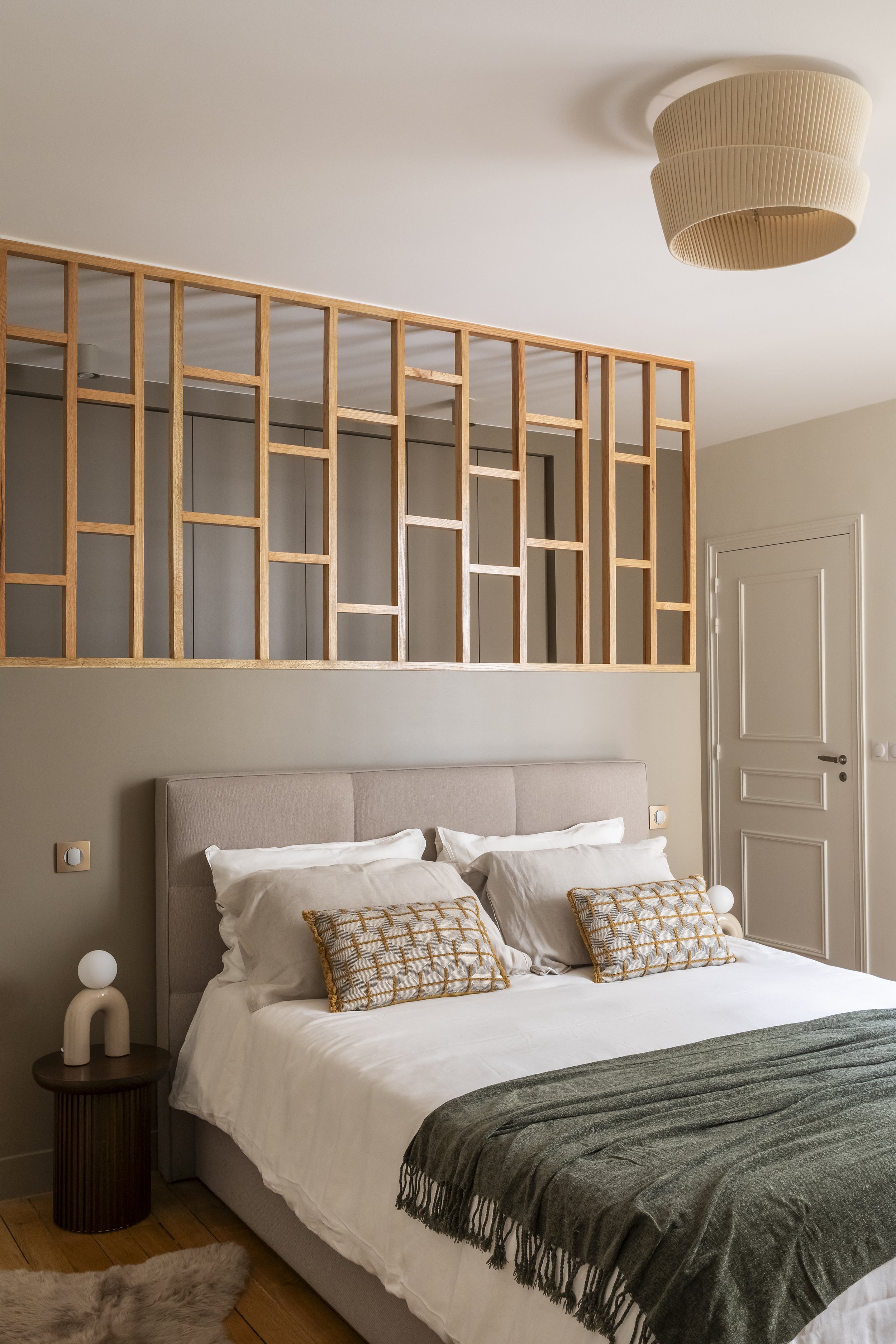 Chambre moderne avec un lit blanc à tête rembourrée, décorée de coussins à motifs dorés, un plaid gris et une lampe de chevet blanche. Un panneau en bois et une porte blanche sont visibles sur le mur.