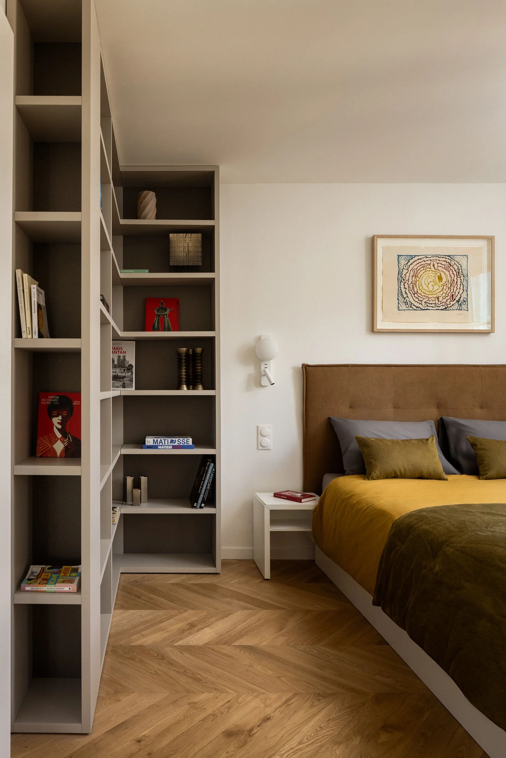 Une chambre parentale dans un intérieur Haussamannien avec un lit en bois avec un boutis couvre-lit en velours de couleur Bronze de la marque Bonsoirs Un tableau À côté du lit, une petite table blanche avec un livre rouge dessus. Le mur est blanc et 