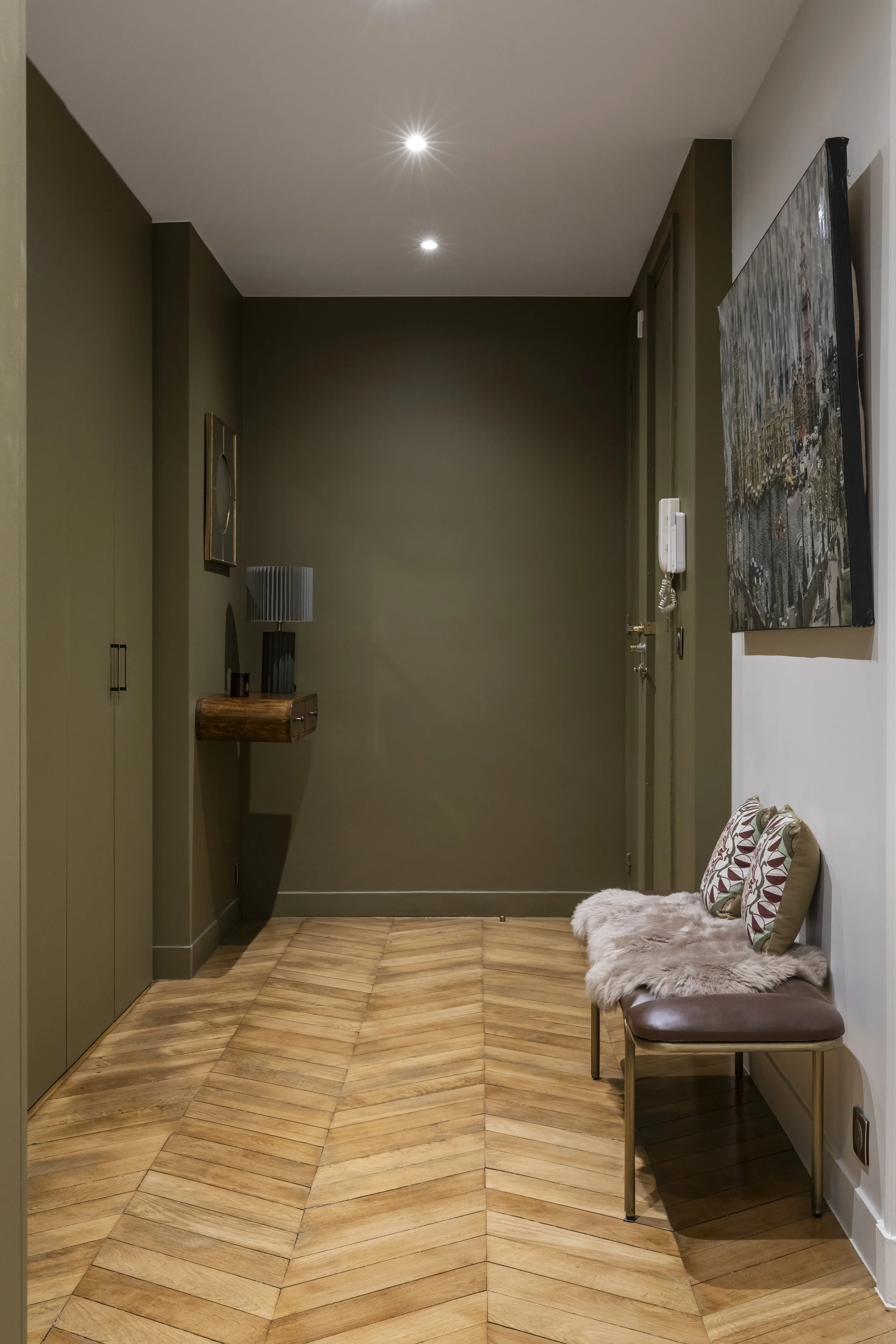 Un couloir dans un appartement moderne avec un sol en parquet en chevron, un mur vert olive, un fauteuil avec coussins et une fourrure, des tableaux et une lampe.