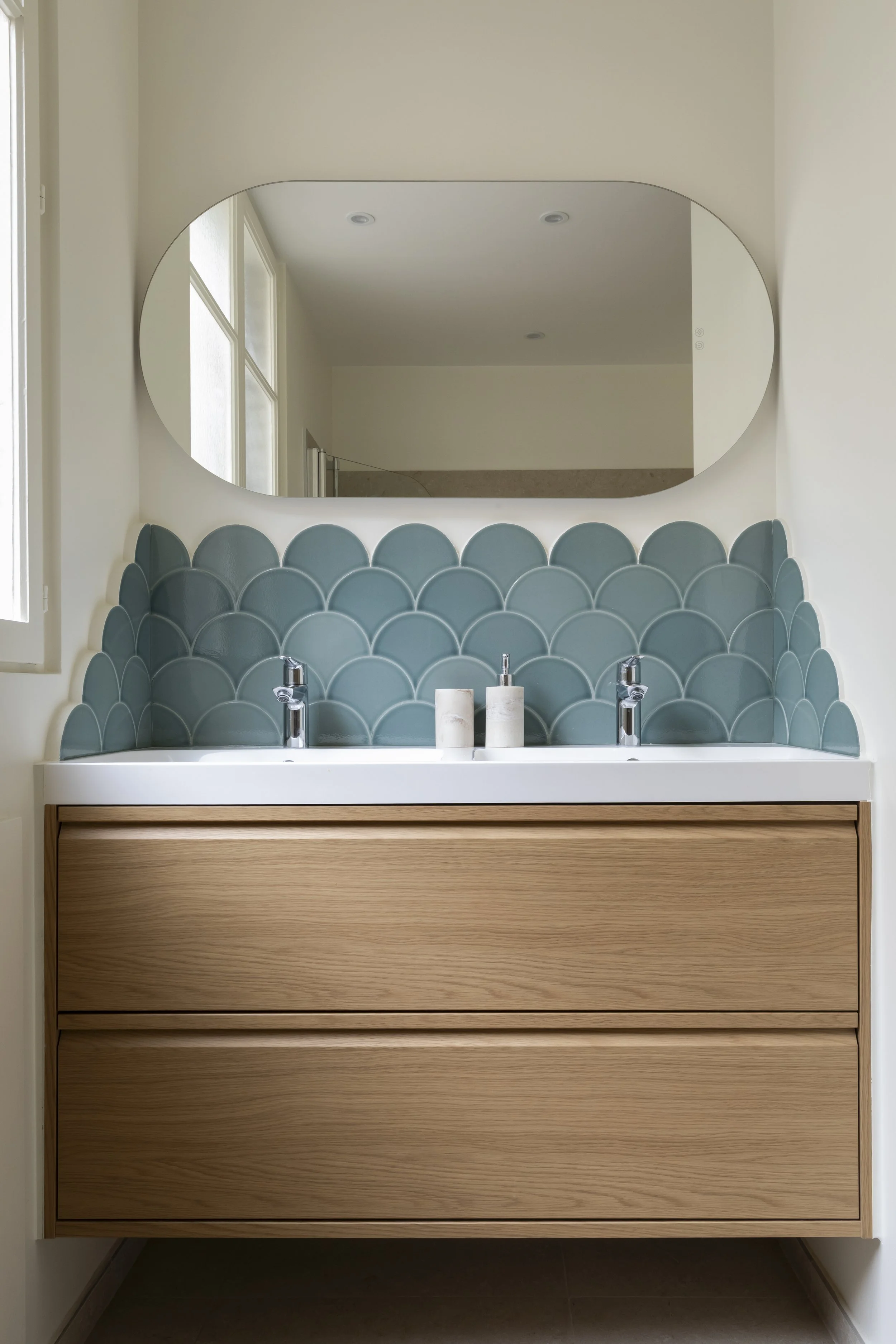 Grand miroir ovale au-dessus d'un lavabo avec mur de carreaux en forme d'écailles bleues, deux pommels de savon blanc, miroir dans une salle de bain lumineuse.
