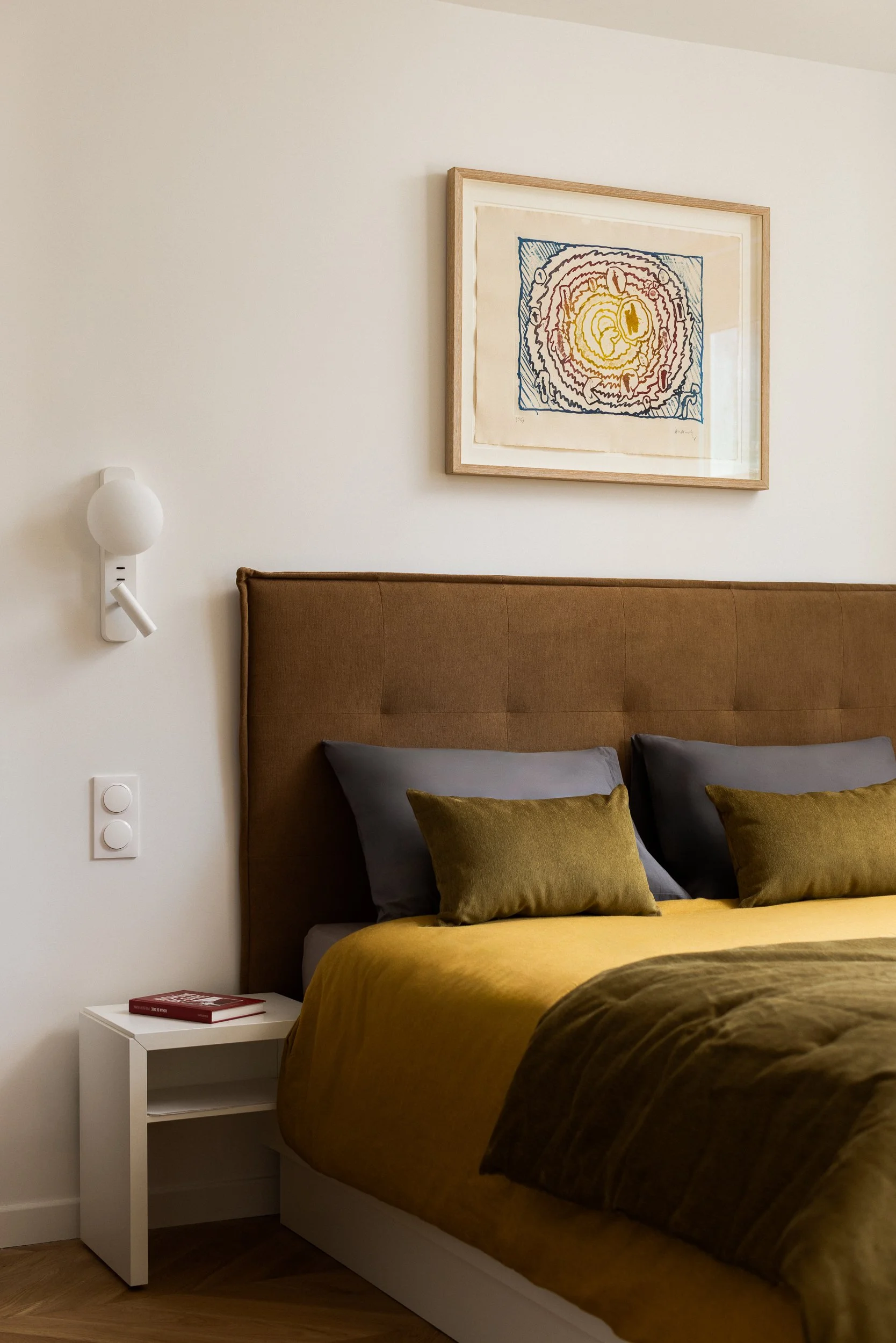 Une chambre parentale dans un intérieur Haussamannien avec un lit en bois avec un boutis couvre-lit en velours de couleur Bronze de la marque Bonsoirs Un tableau À côté du lit, une petite table blanche avec un livre rouge dessus. Le mur est blanc et 