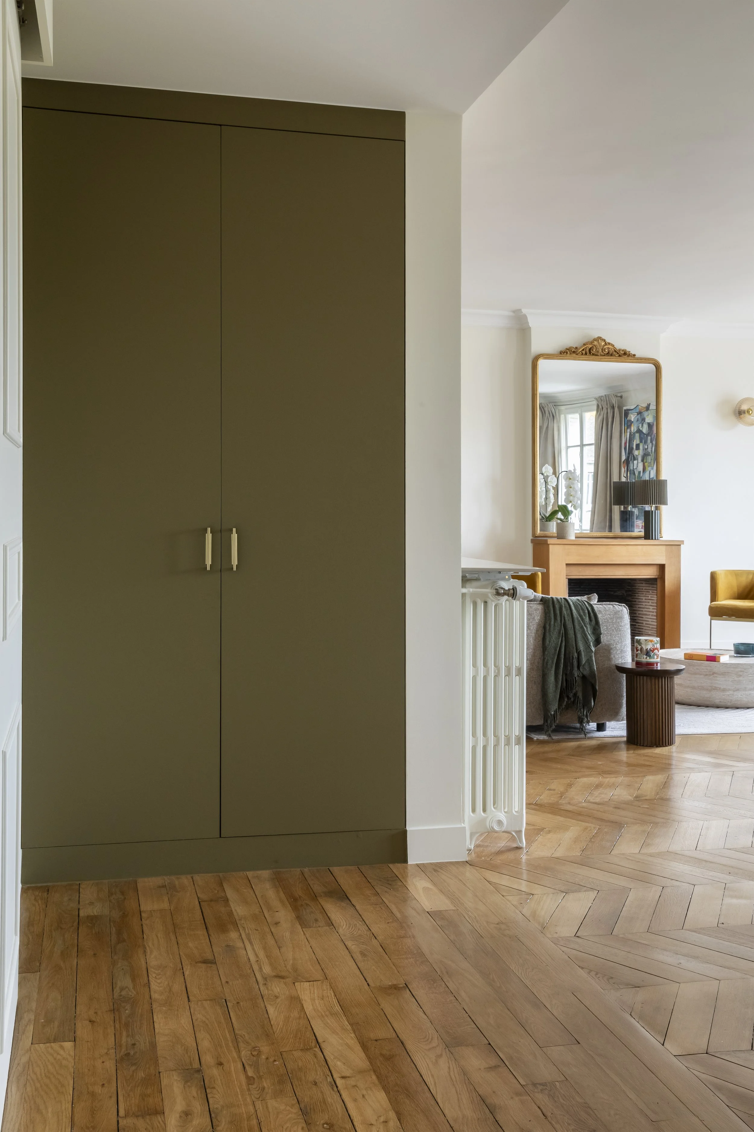 Intérieur d'une pièce d'appartement avec parquet en bois, un mur beige, un grand miroir avec cadre doré au dessus d'une cheminée en brique, un canapé gris avec une couverture verte, un petit tabouret en bois avec une tasse, et une table basse.