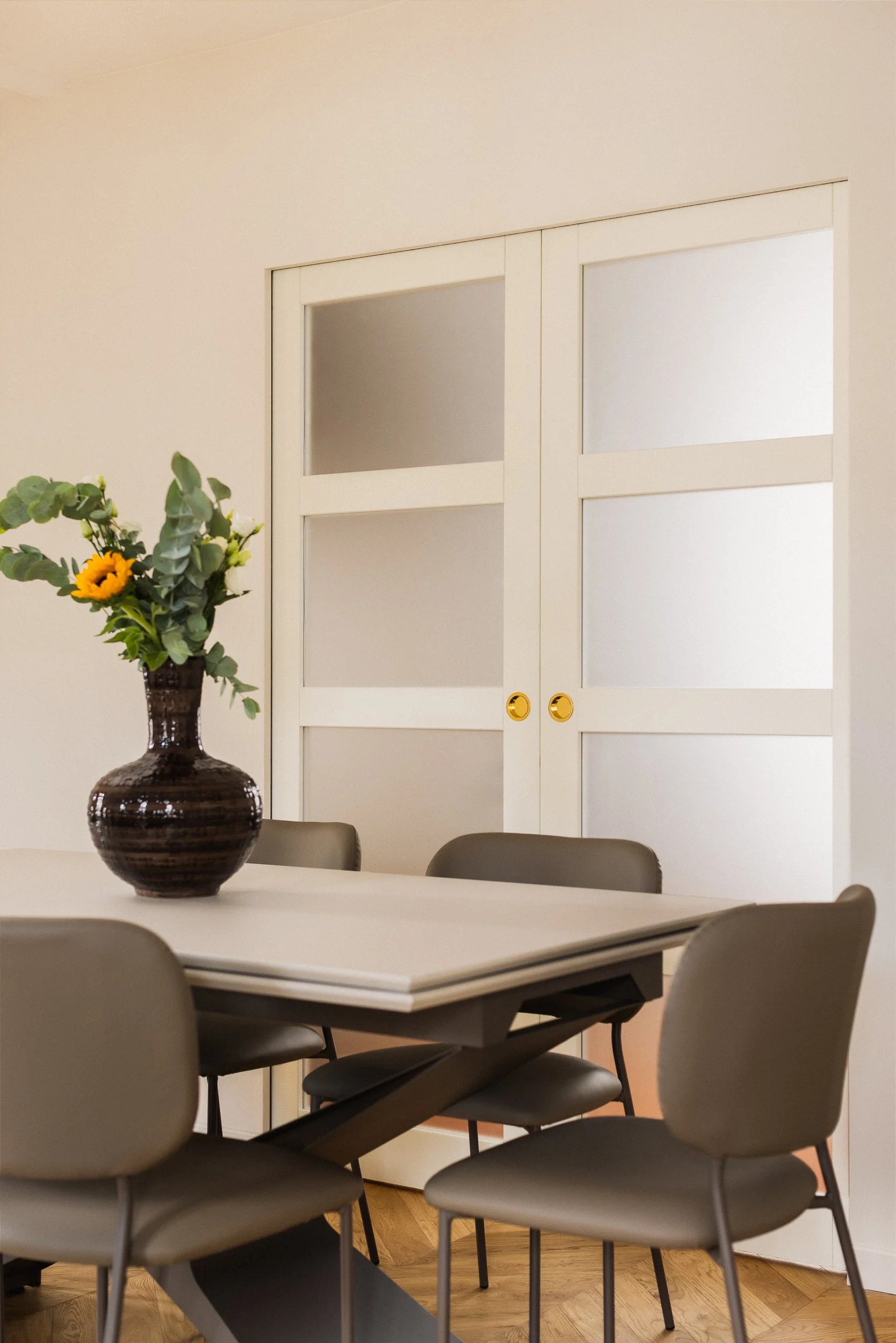 Une salle à manger contemporaine dans un intérieur Haussmannien avec une table en métal extensible entourée de 6 chaises en métal et cuir. Au centre de la table, un vase noir avec des fleurs vertes et une fleur jaune. À l'arrière une porte à galandag