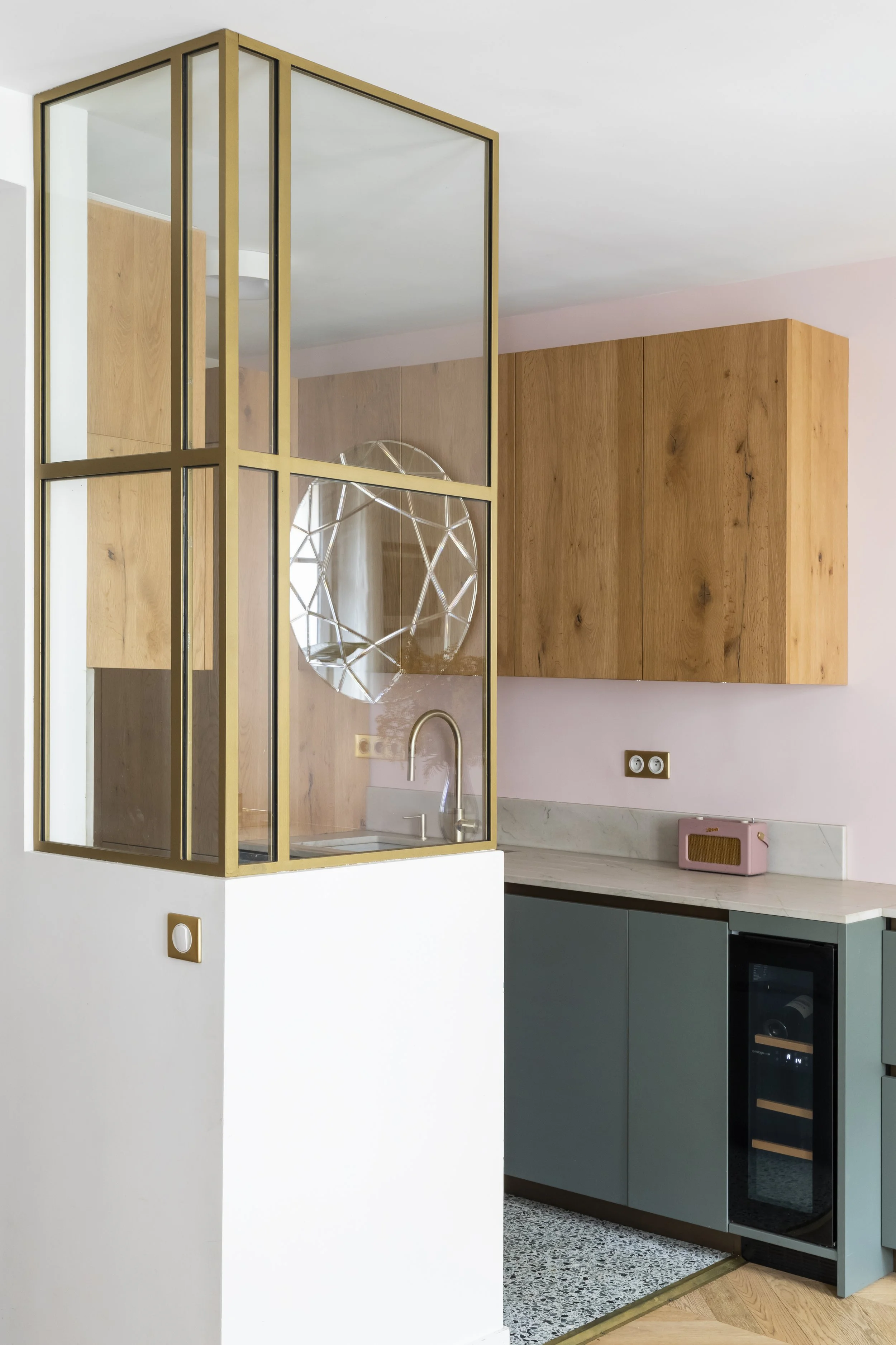 Cuisine moderne avec un meuble en bois, un four occulté par des portes bleues, un mur en bois clair, un plan de travail en marbre, et une cloison en verre avec une structure métallique dorée.