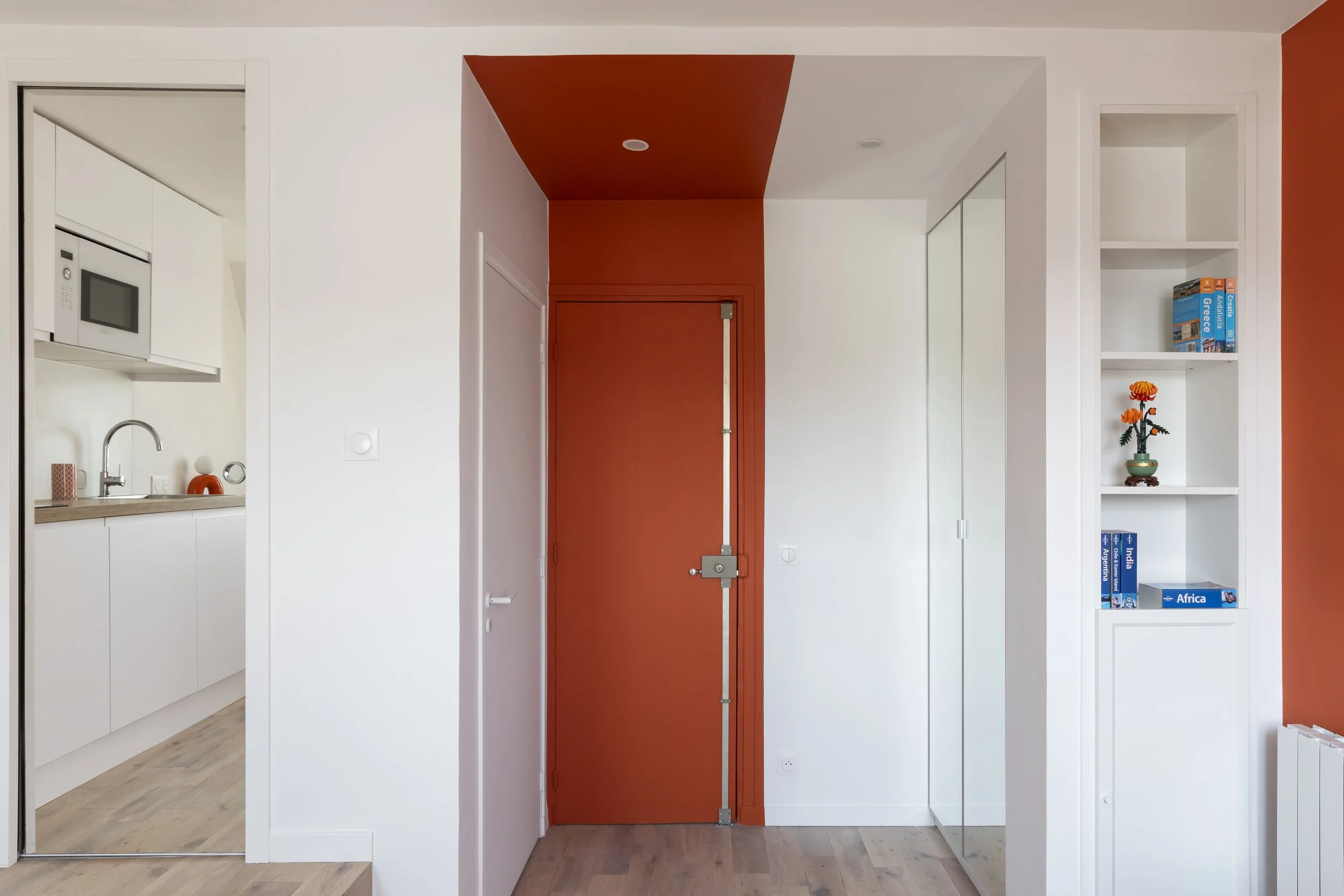 Un mur intérieur blanc avec une porte en métal orange au centre, entouré de deux murs blancs, un avec une étagère blanche contenant des livres sur l'Union africaine et une petite décoration. Un coin cuisine avec un évier, un micro-ondes et quelques a