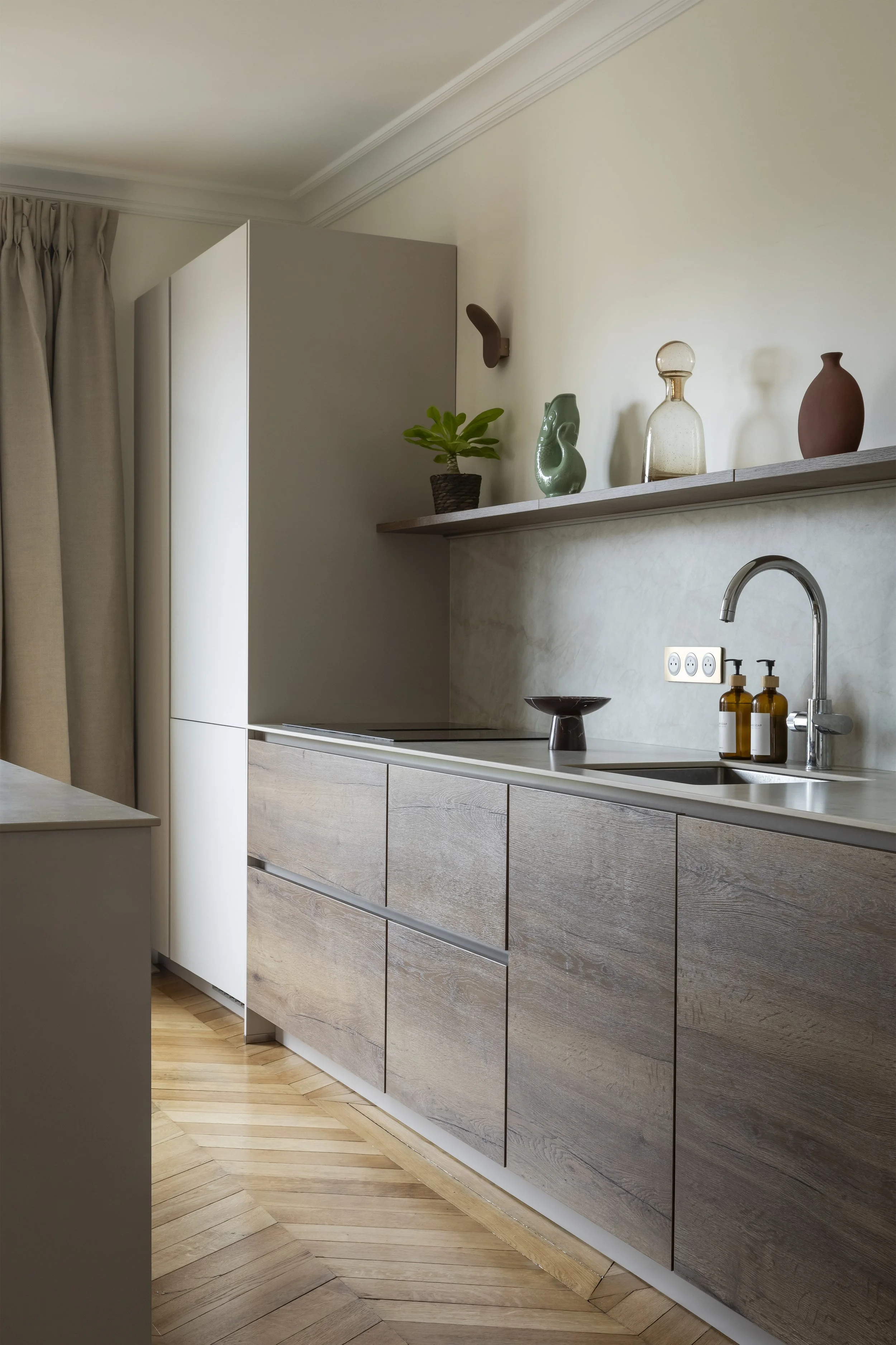 Cuisine moderne contemporaine ouverte sur pièce à vivre salon avec îlot central avec un plan de travail en céramique, des armoires et  en bois clair, une étagère avec décorations. La pièce est lumineuse avec des murs beige et parquets en bois clair p