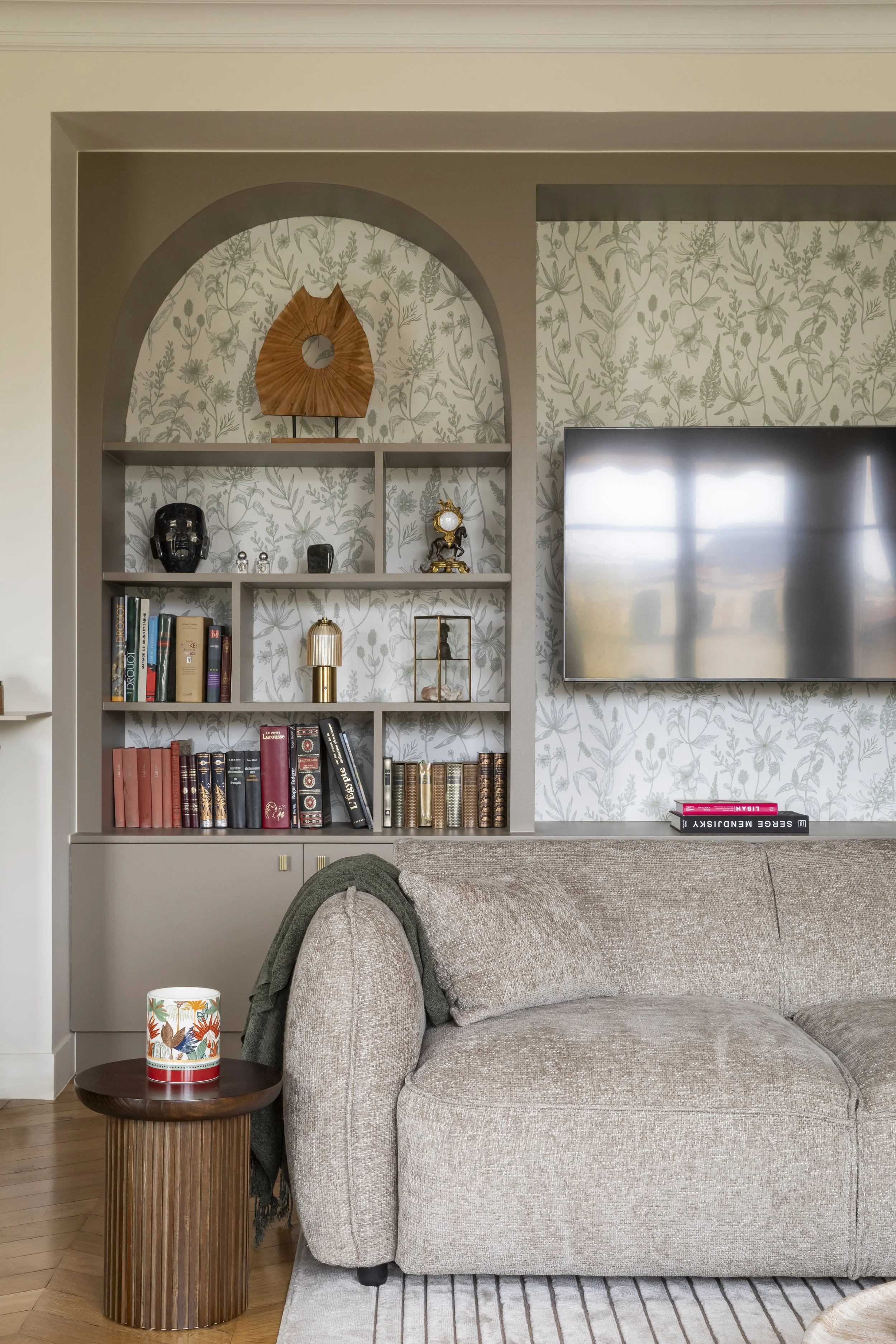 Salon avec étagères décoratives contenant des livres, des objets d'art et une horloge. Une télévision est fixée au mur, et un canapé beige est placé devant avec un tapis rayé au sol.