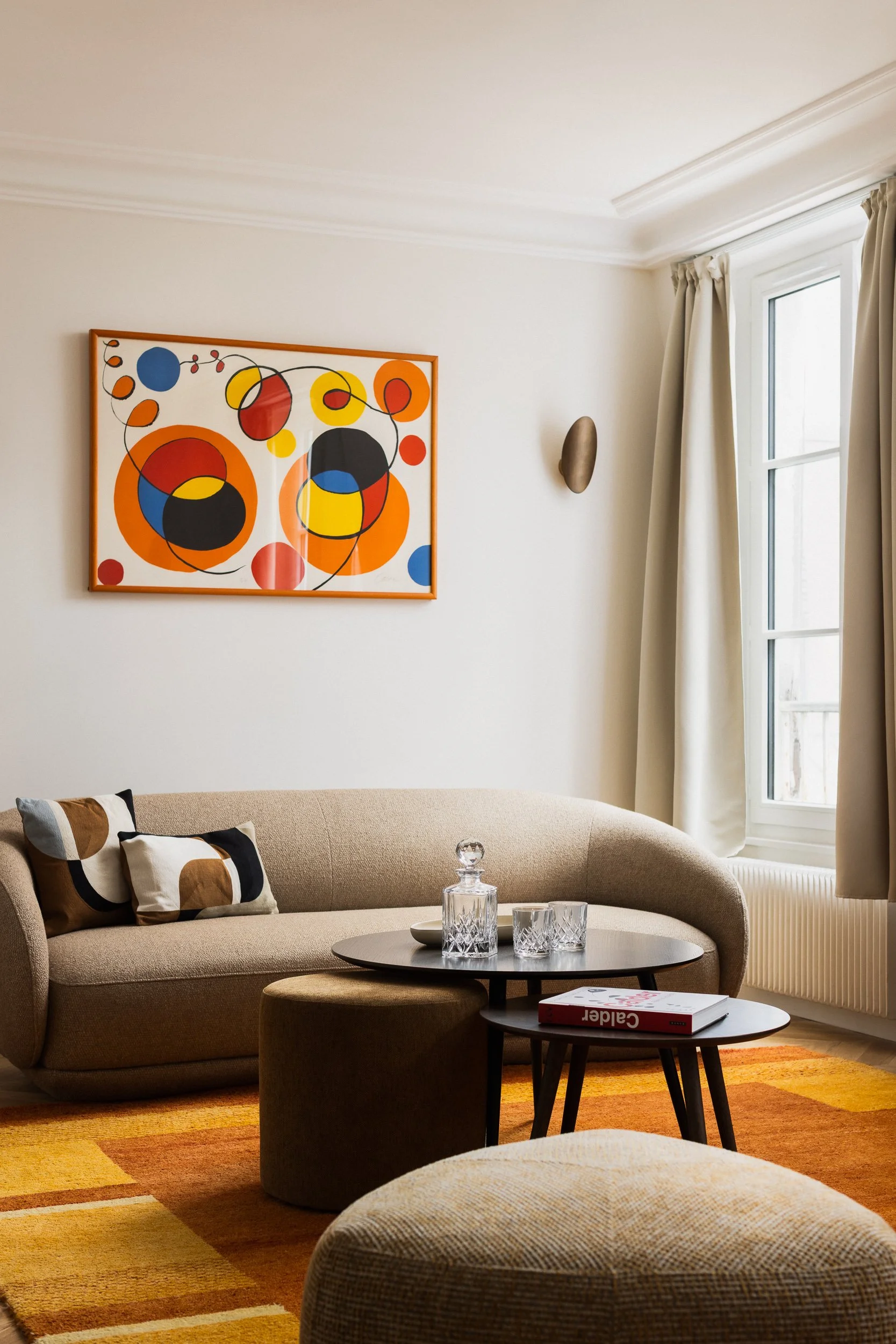 Salon contemporain dans appartement haussmannien avec canapé beige arrondi de la marque Boconcept modèle Bolsano, pouf triangulaire aux angles arrondis de la marque Boconcept modèle Bermuda, tableau abstrait coloré sur le mur, tables basses rondes gi