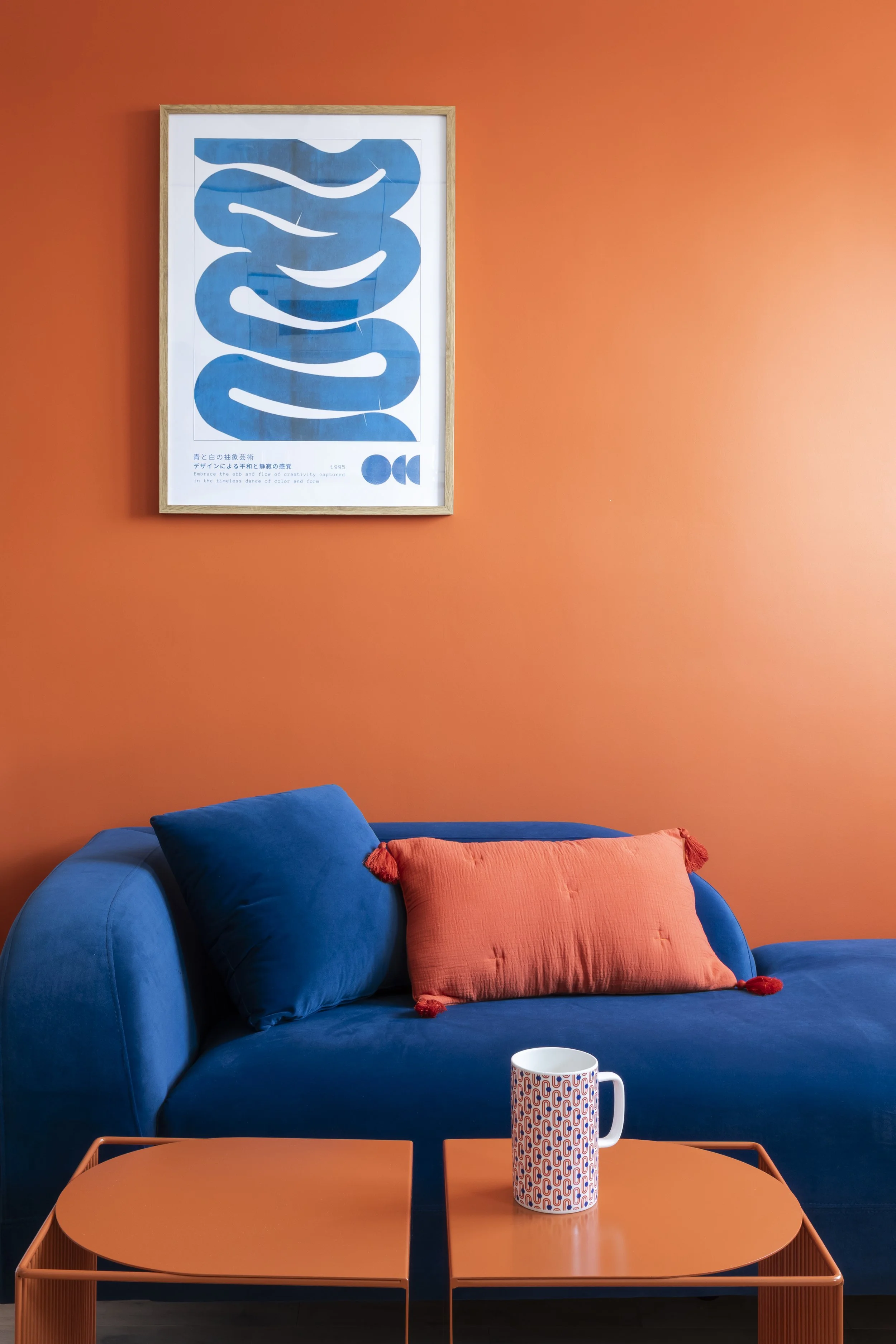 Un canapé bleu avec deux coussins, un orange et un bleu, devant un mur orange. Une table orange avec une tasse délicate à motifs colorés et une œuvre d'art abstraite bleue dans un cadre en bois est suspendue au mur.