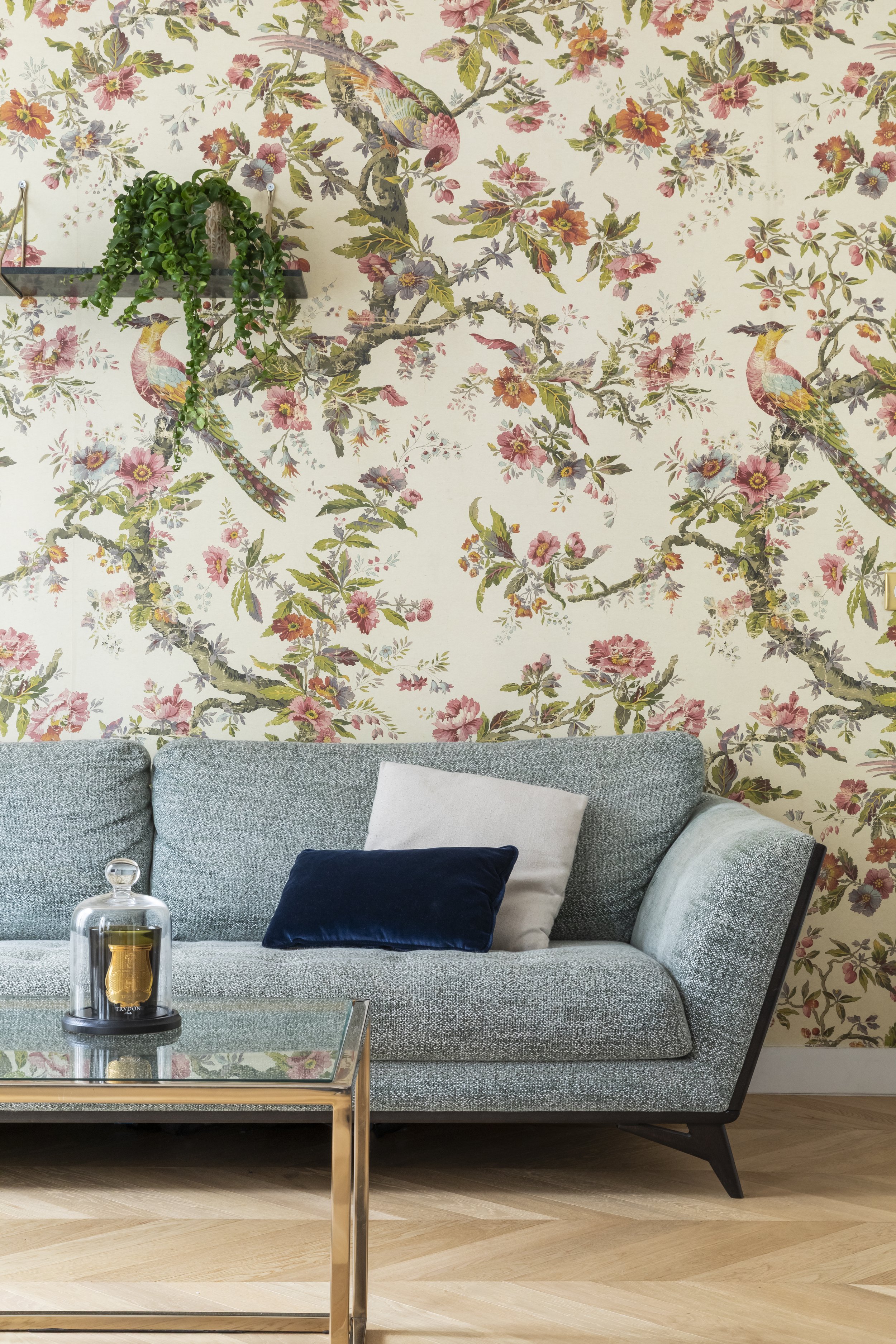 Un salon avec un canapé gris, plusieurs coussins, une table en verre avec une bougie, un mur avec un papier peint floral avec des oiseaux, et une plante en suspension.
