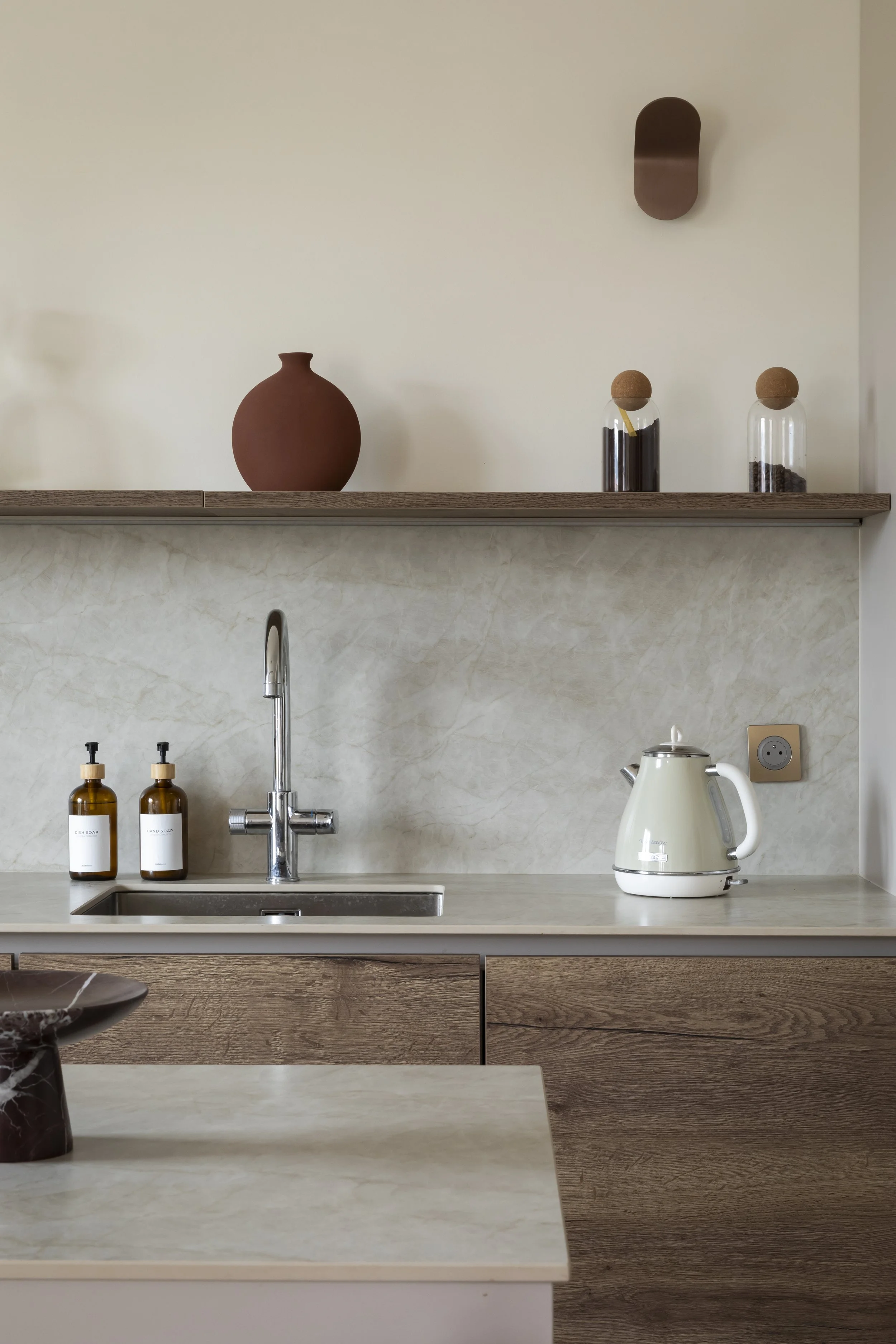 Cuisine moderne contemporaine ouverte sur pièce à vivre salon avec îlot central avec un plan de travail en céramique, des armoires et  en bois clair, une étagère avec décorations. La pièce est lumineuse avec des murs beige et parquets en bois clair p